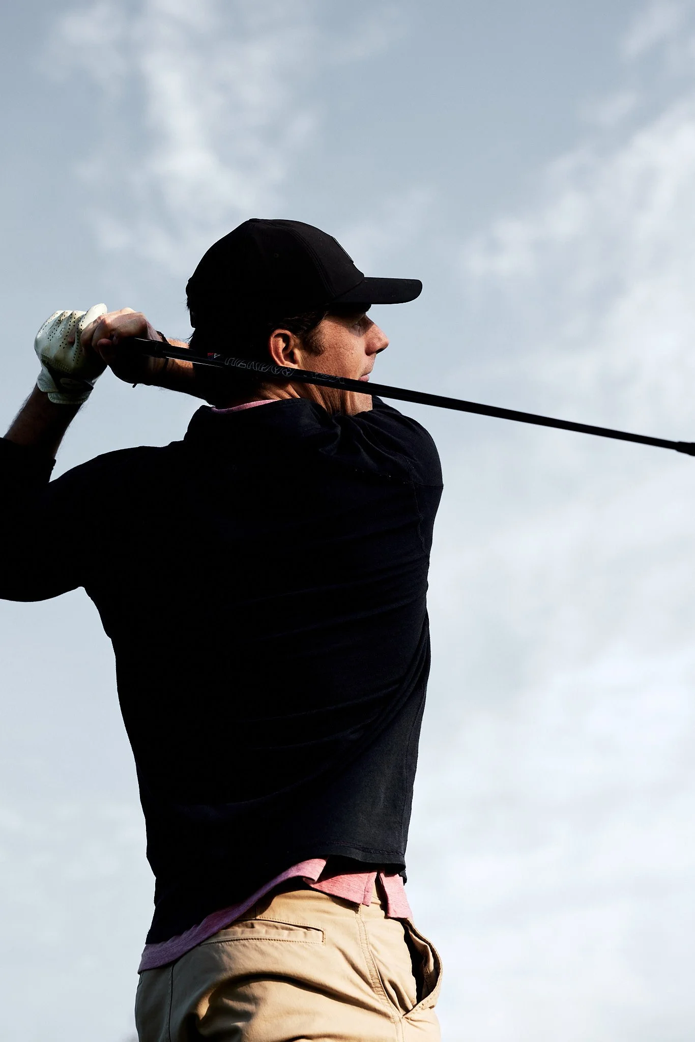 A man wearing a black cap, black shirt, and beige pants is swinging a golf club against a cloudy sky.