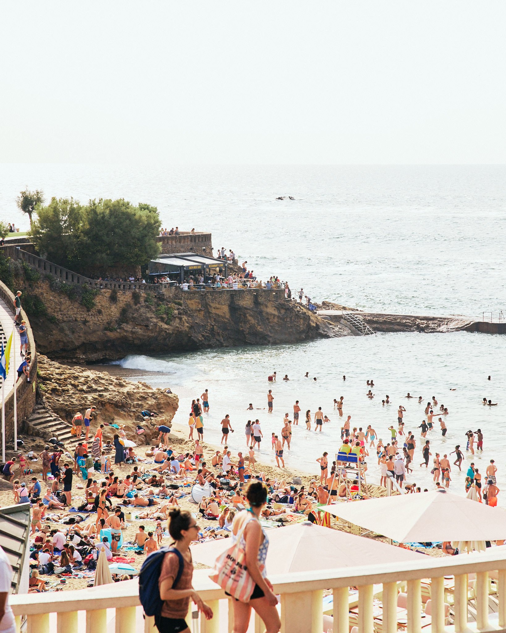 Crowded beach scene with many people sunbathing on sand, swimming, and walking along the shoreline under umbrellas and in the water. The background shows cliffs with an outdoor cafe and people sitting on a deck overlooking the ocean.