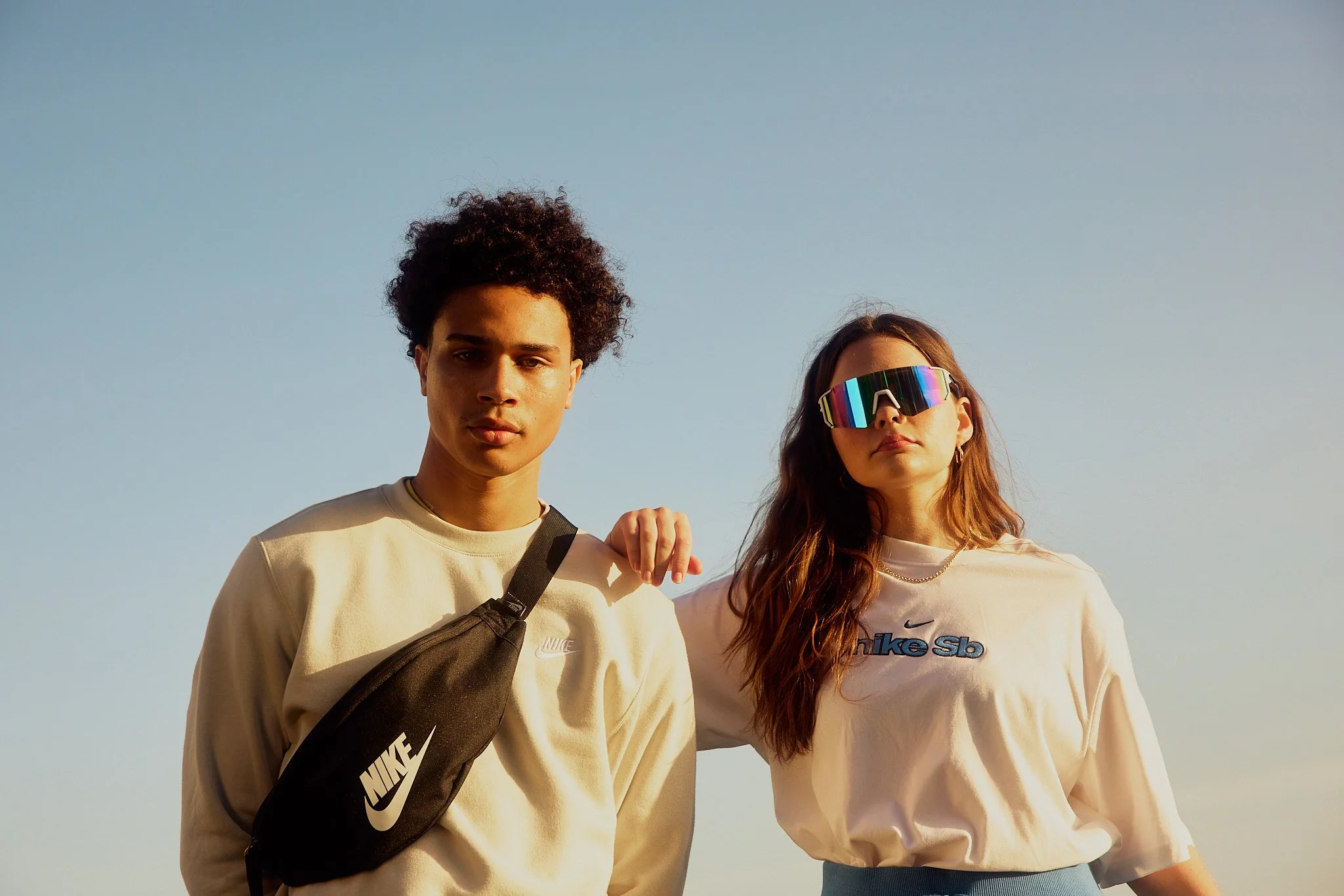 A young man and woman stand outdoors against a clear blue sky, with the young woman wearing reflective sunglasses and the young man carrying a Nike shoulder bag.