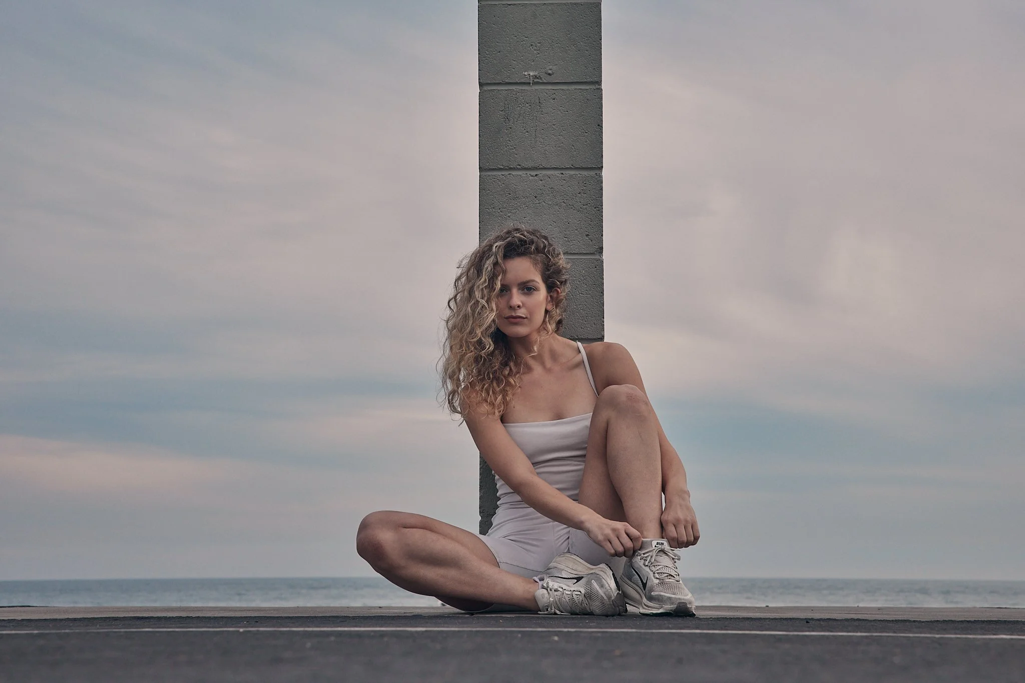 A woman with curly hair, wearing a light-colored tank top and shorts, is sitting cross-legged on a wooden surface near the beach. She is leaning against a concrete post, with a cloudy sky and the ocean in the background.