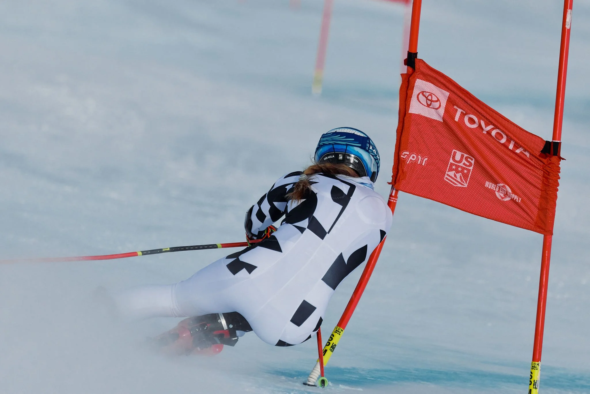 An athlete in a blue helmet and black and white racing suit slides on snow during a skiing race, navigating around red and yellow gate flags.
