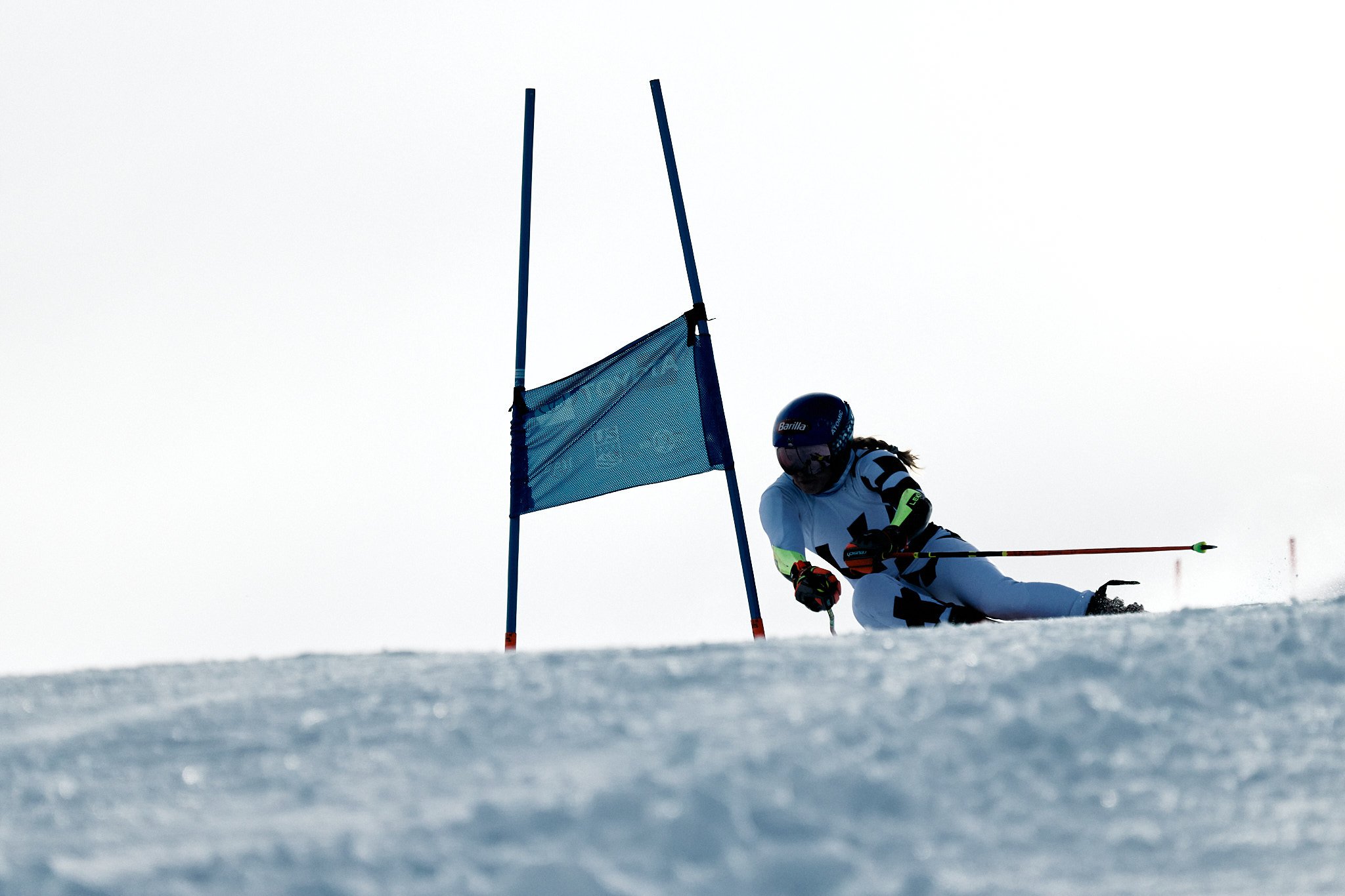 A skier racing downhill on a snowy slope, leaning into a turn with a blue flag marking the course, in an overcast or foggy sky.