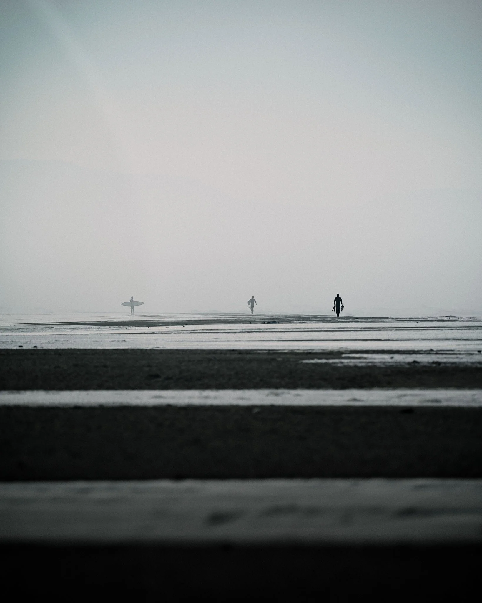 Silhouettes of three people walking along a foggy beach with a person holding a surfboard in the distance.