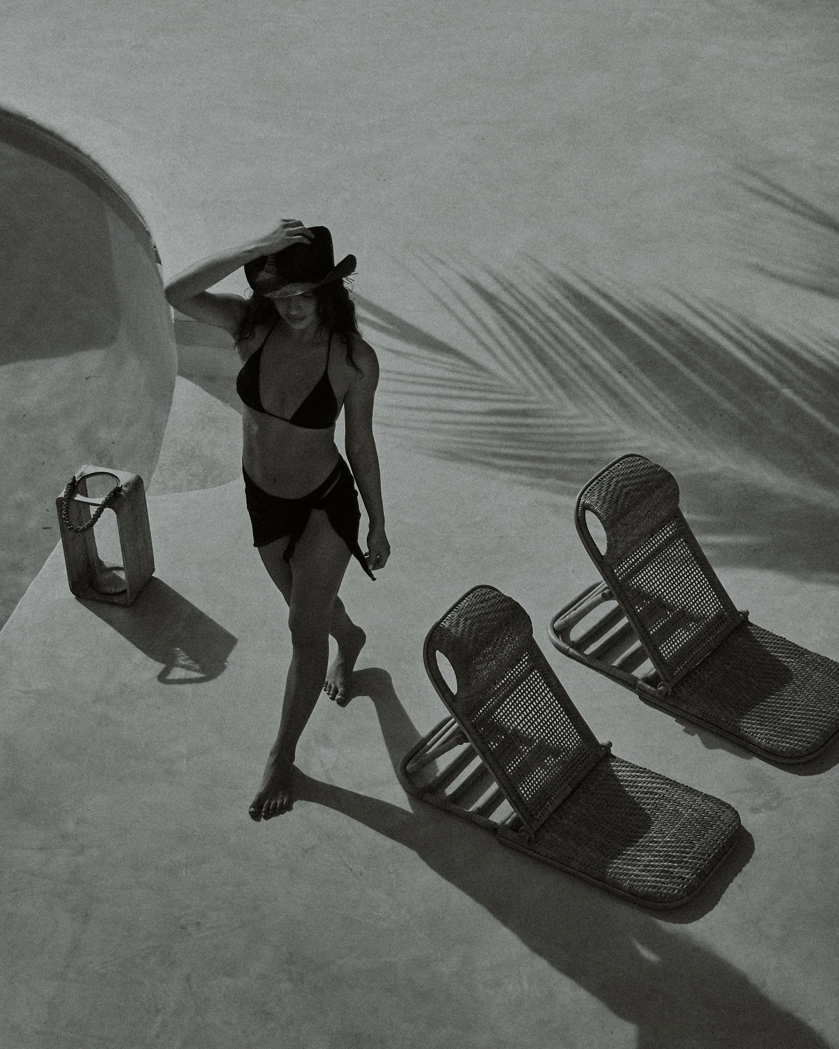 A woman in a bikini and wide-brimmed hat standing on a beach with a small table, two chairs, and shadows cast on the sand.