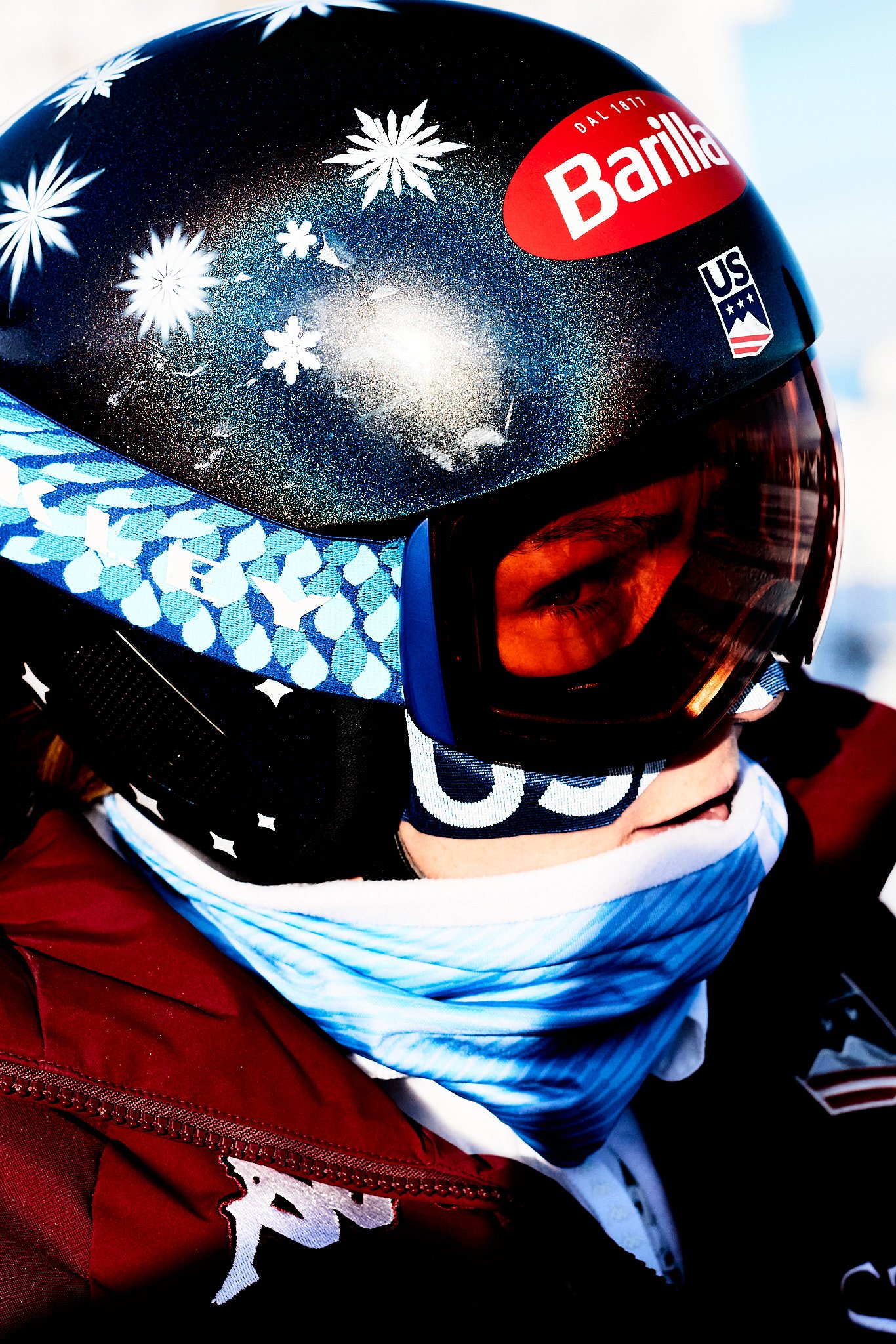 Close-up of a person wearing a black helmet with white snowflake patterns, a red sticker with 'Barilla' logo, and ski goggles. The person also has a white neck gaiter and red jacket, with part of a blue and white patterned face mask visible.