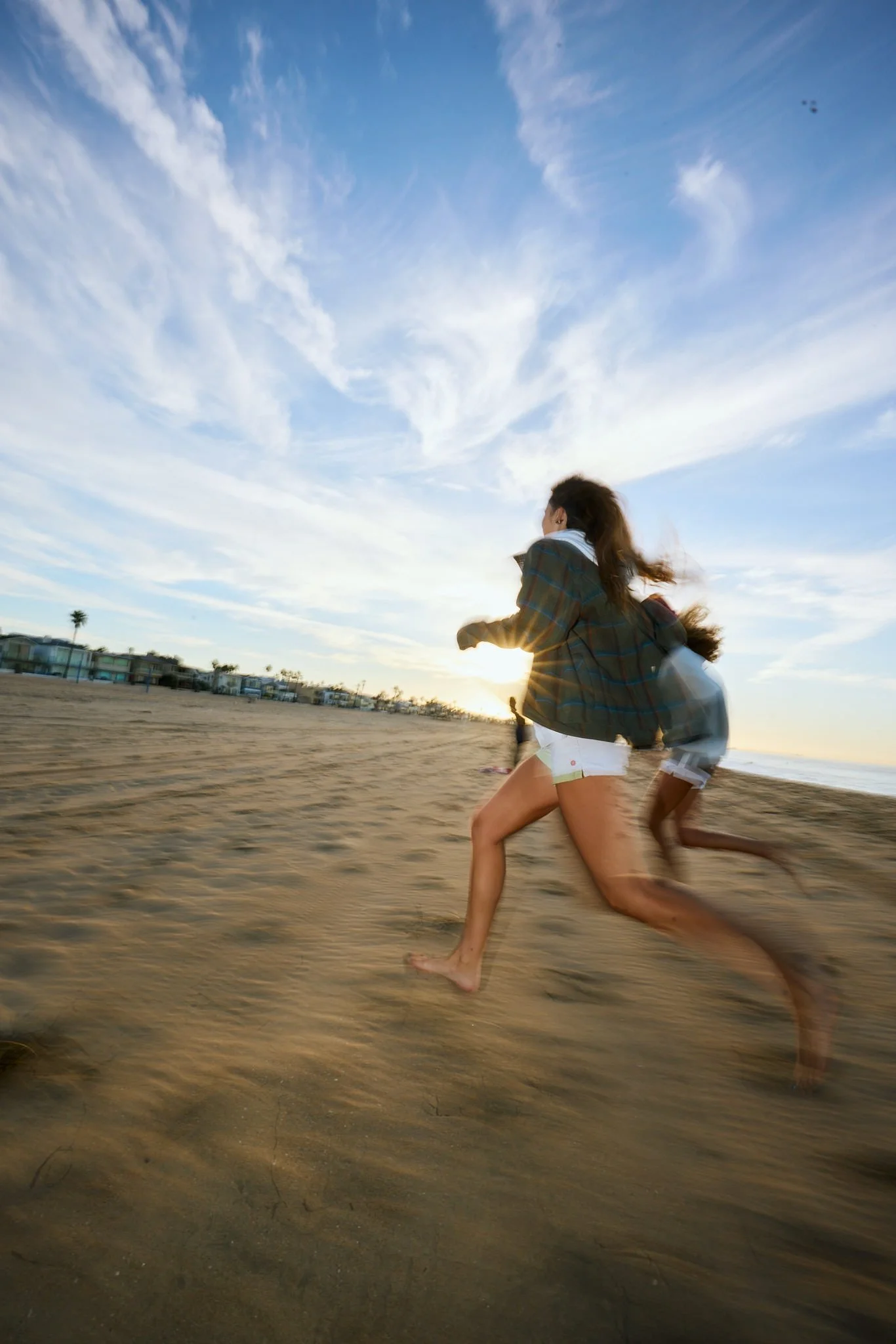 People running on a sandy beach at sunset, with a partly cloudy sky and buildings in the background.