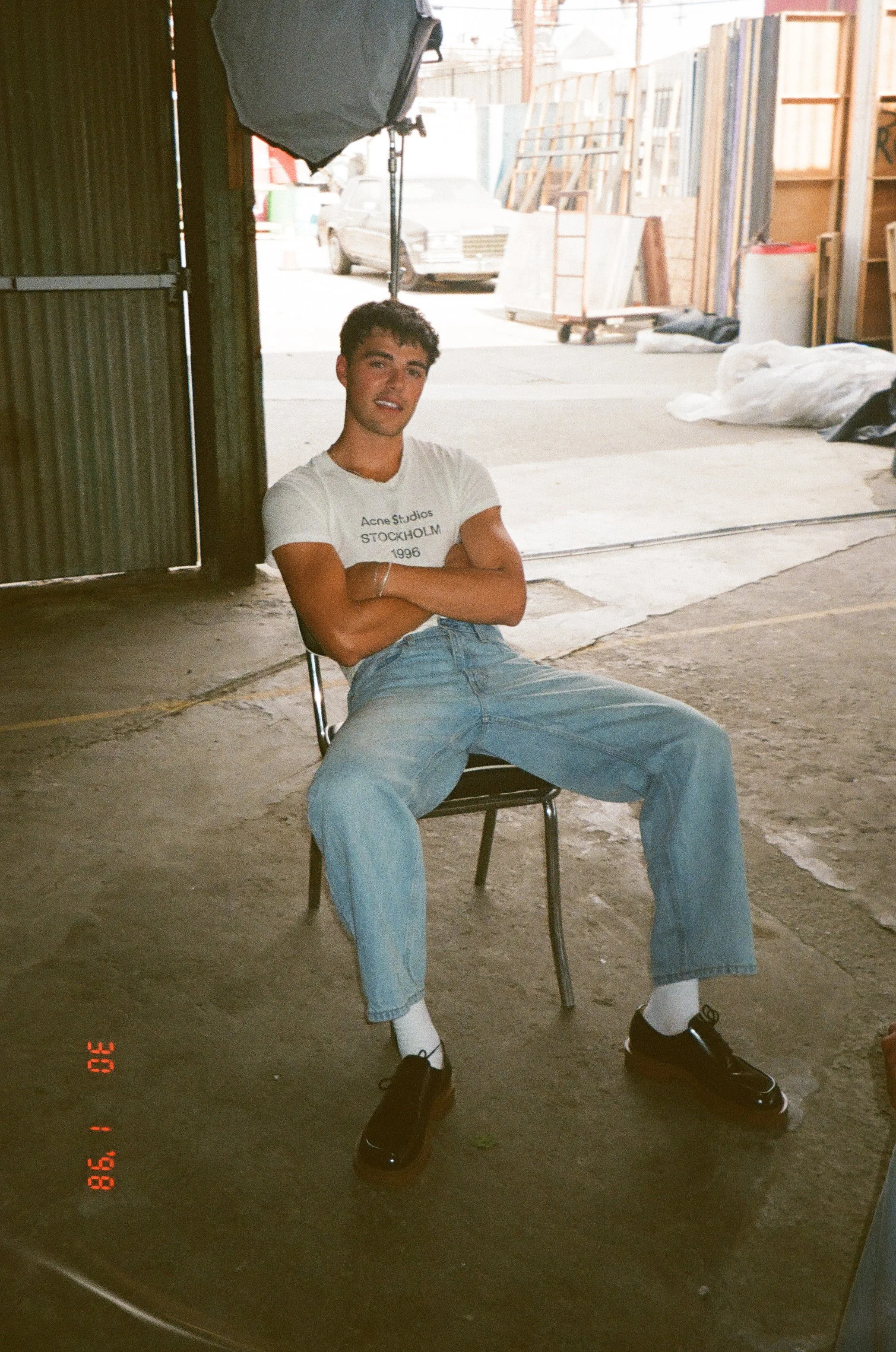 Young man sitting on a chair in an industrial workshop or warehouse, with wooden and metal materials visible in the background, wearing a white t-shirt and jeans.