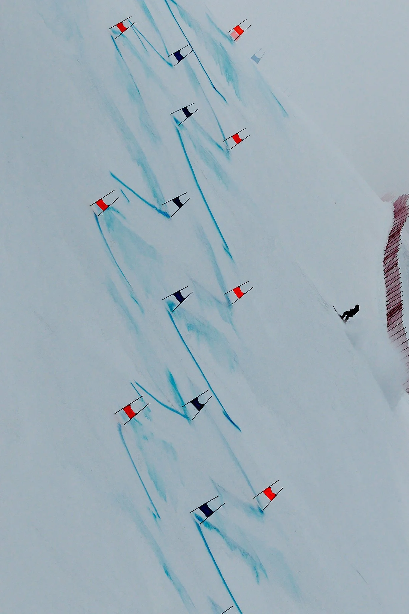 A skier racing down a snow-covered slope, marked with multiple flags, with blue ski tracks in the snow and a skier wearing dark clothing at the bottom right.