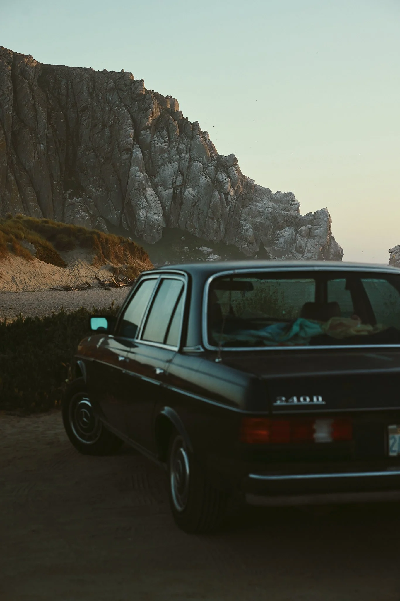 A black Subaru car parked near a coastal area with large rocky cliffs in the background during sunset or dusk.