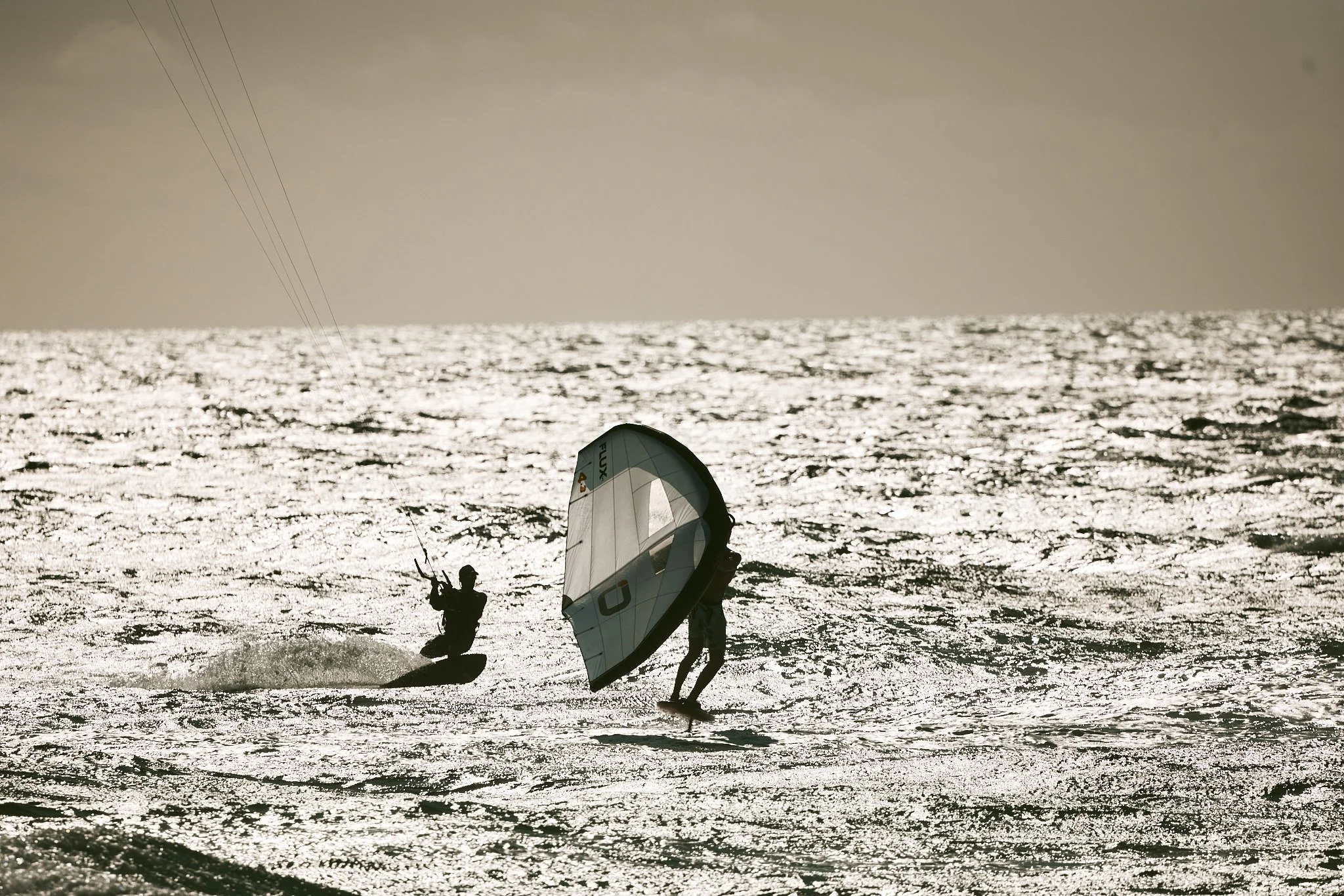 Two people windsurfing on the ocean during sunset with a bright, reflective surface and a dark sky.