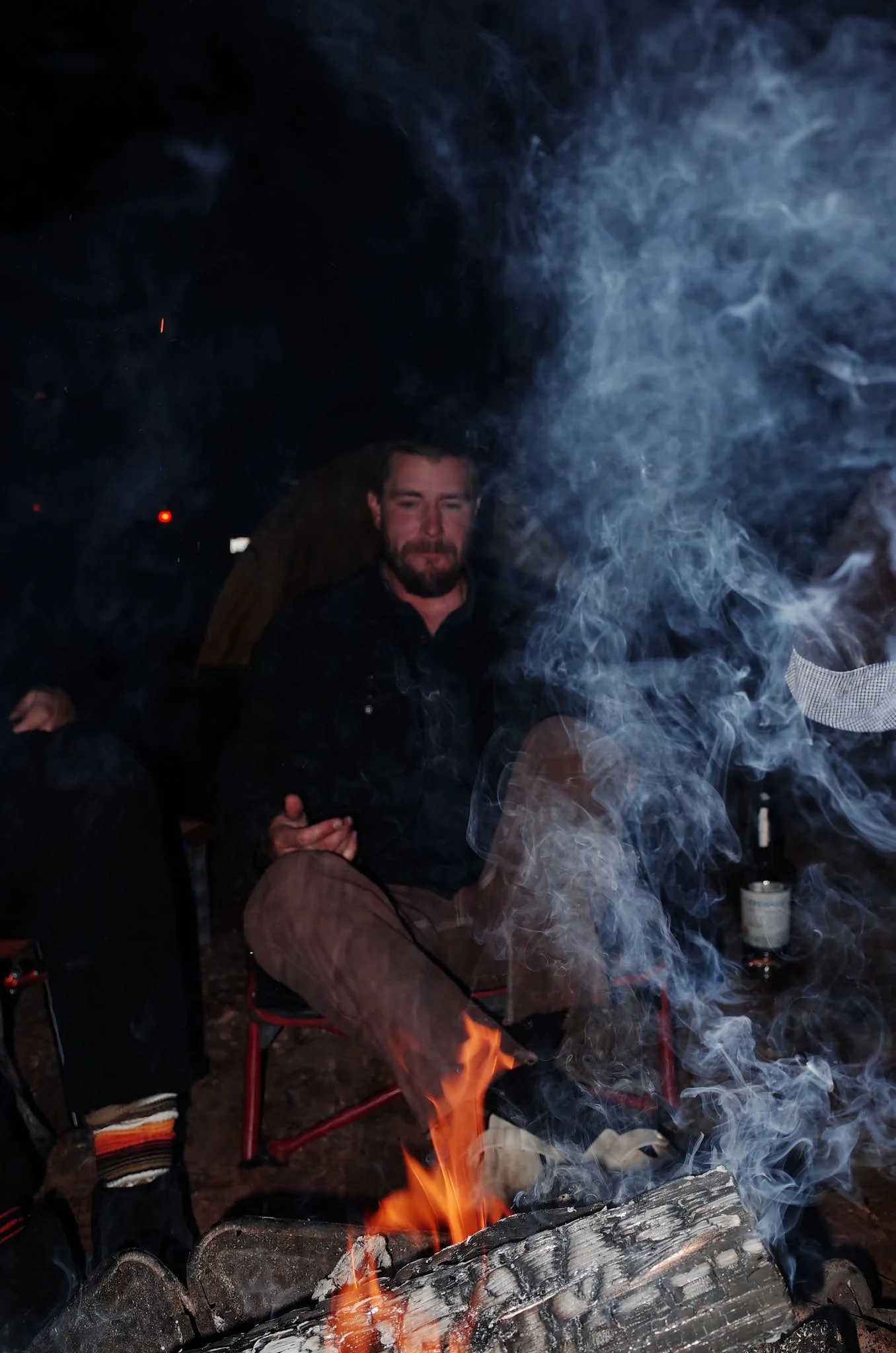 A man sitting beside a fire with smoke around him, holding a glass, at night in an outdoor setting.