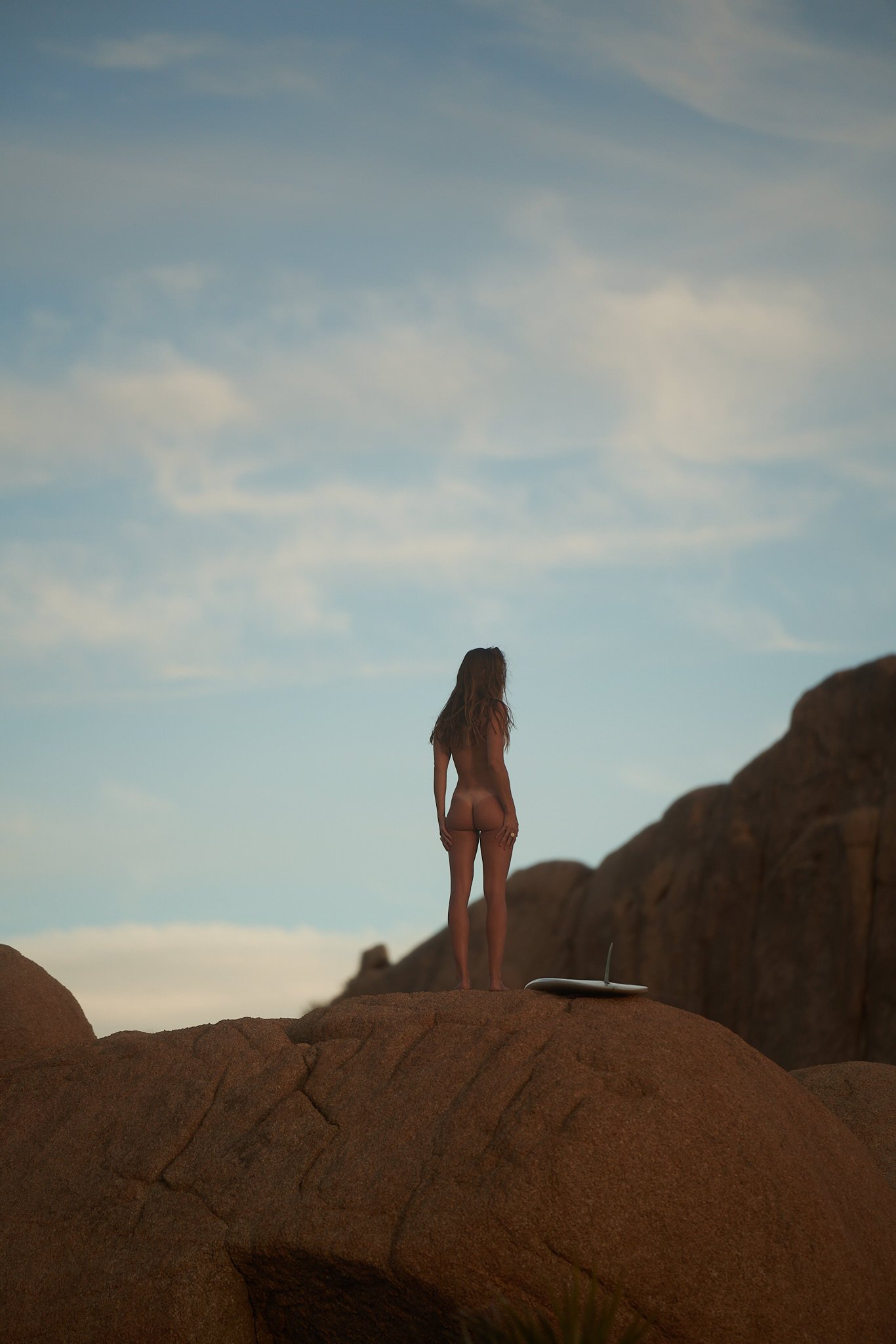 A woman standing on rocks at sunset or dusk, looking out into the distance, with surfboard on the ground nearby.