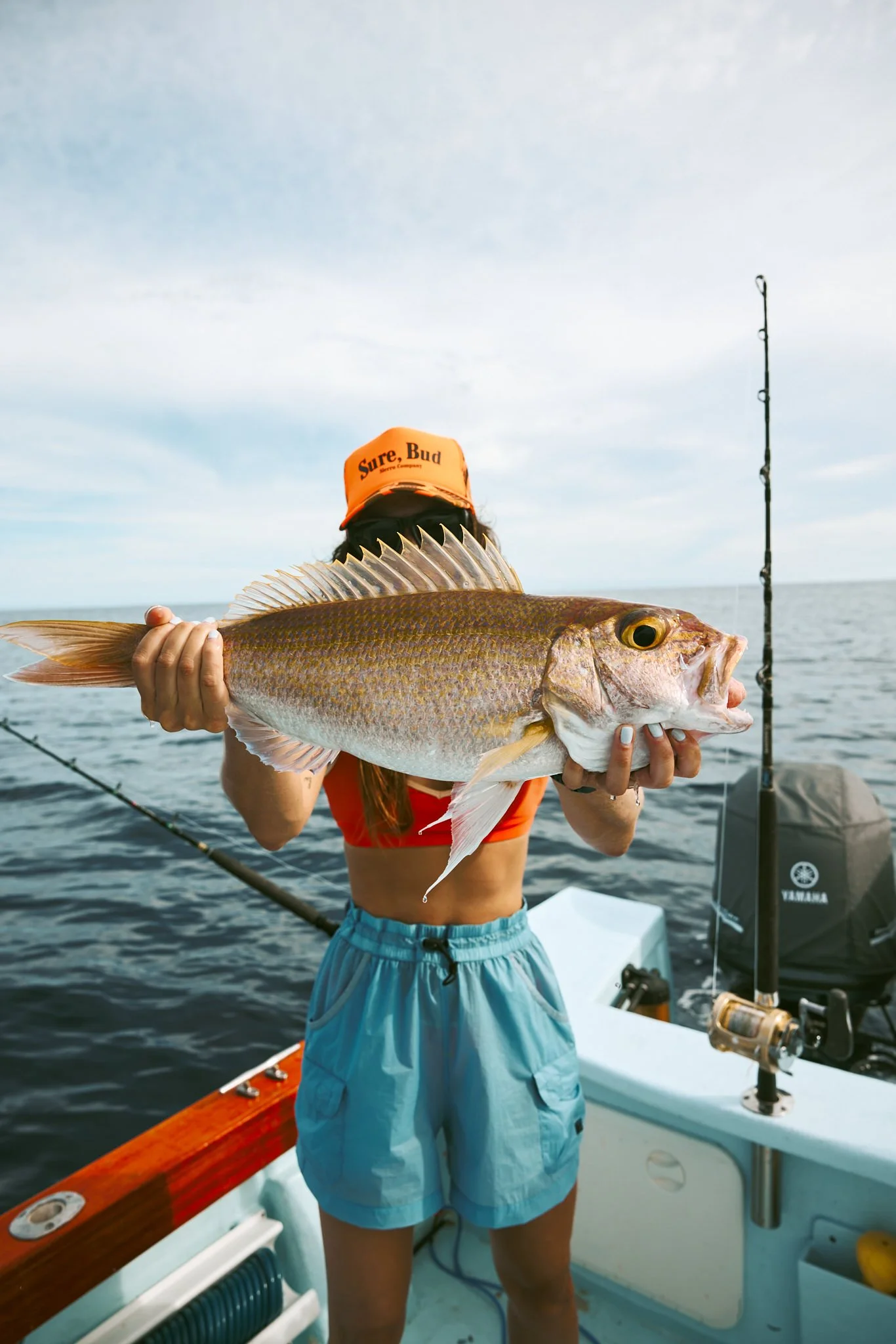 A person holding a large fish on a boat, with a fishing rod in the water and a cloudy sky in the background.