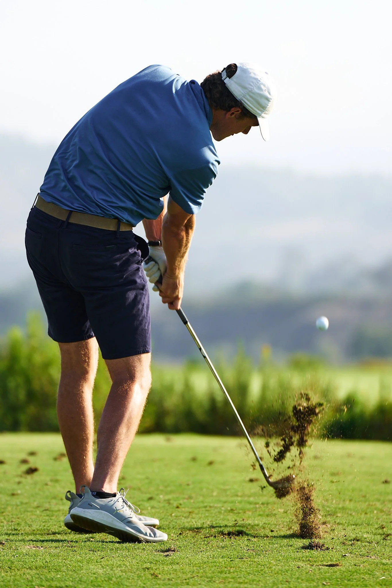A man hitting a golf ball on a golf course, wearing a white cap, blue polo shirt, navy shorts, and white golf shoes.