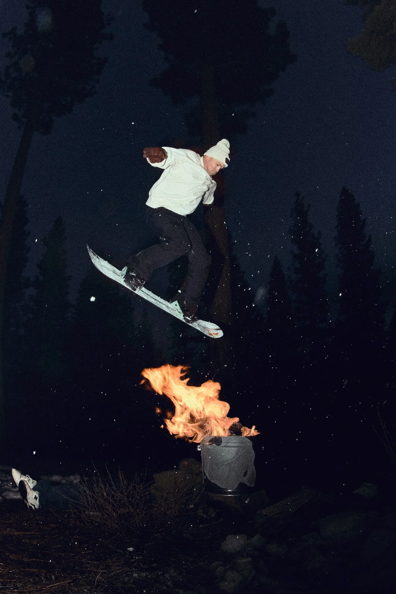 A person dressed in a grey hoodie and pants is sitting in front of a large burning fire at night. The person appears to be looking at the fire, which is producing bright flames. The background features dark trees and a starry sky.