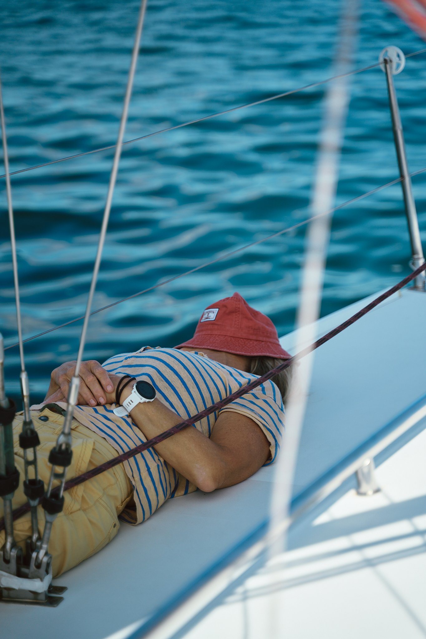 A person lying down on a boat, wearing a red hat, striped shirt, and yellow shorts, with a watch on their wrist, surrounded by boat ropes and water in the background.