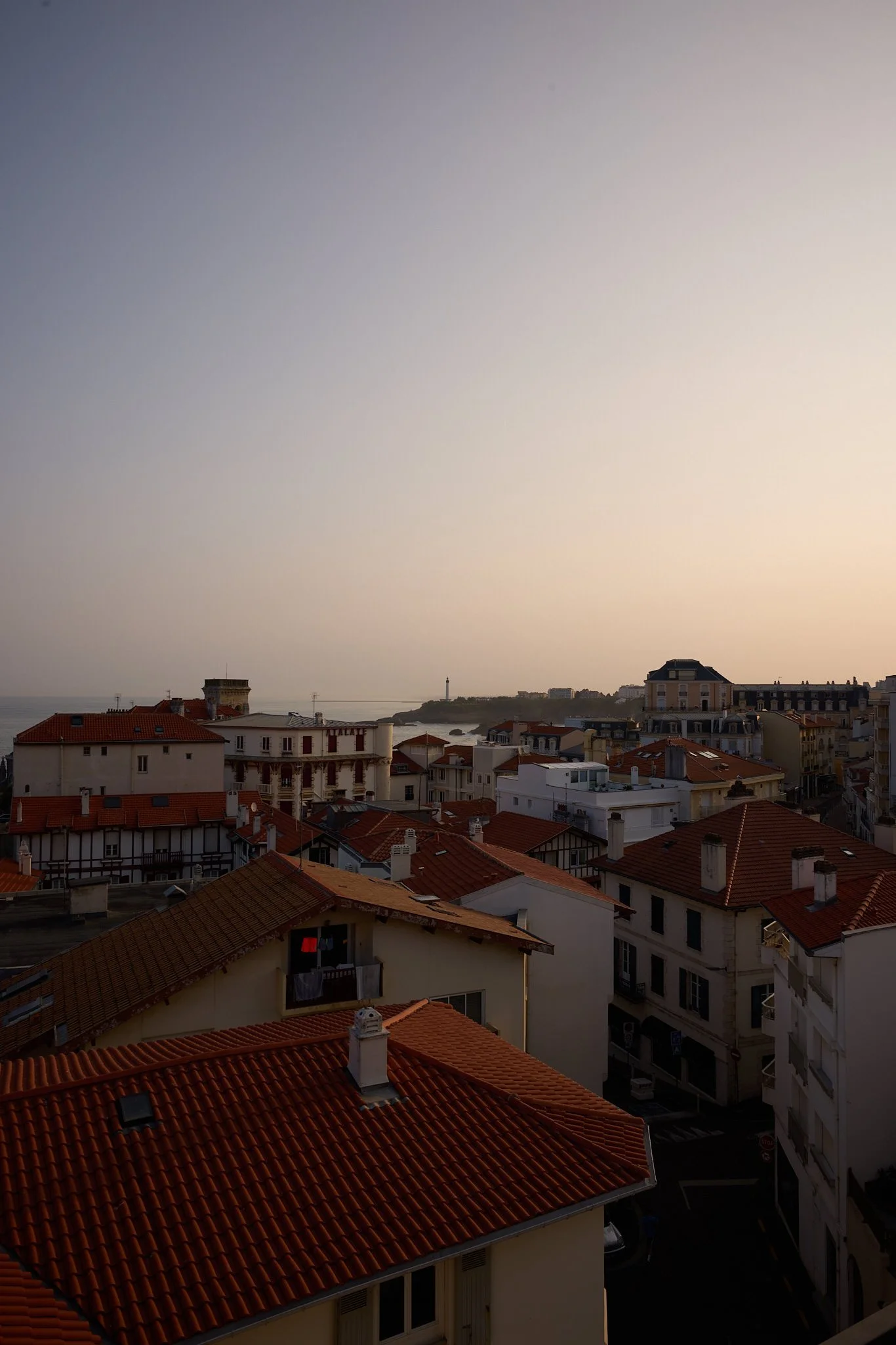 Cityscape with red-tiled rooftops, buildings, and a distant lighthouse under a hazy sky.