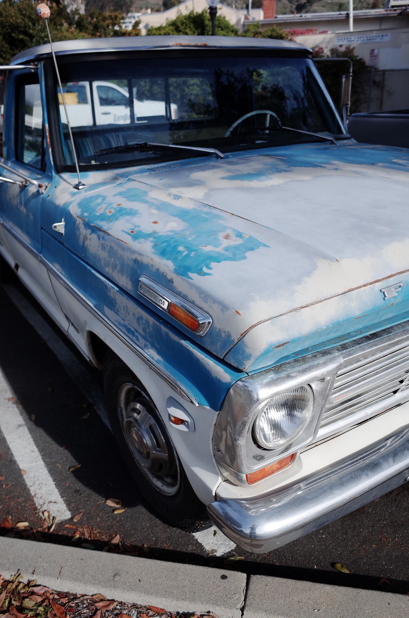A vintage blue and white sedan car with signs of rust and paint chipping, parked in a parking lot with fallen leaves around.