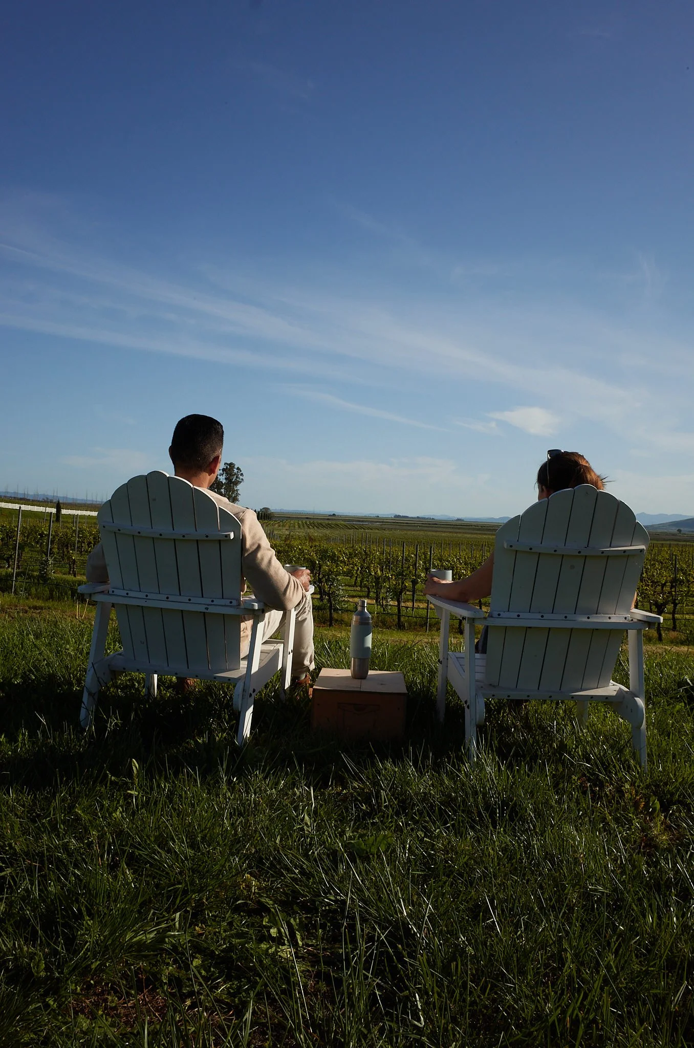Two people sitting in white Adirondack chairs on a grassy field, facing a vineyard and a distant horizon under a blue sky with wispy clouds.