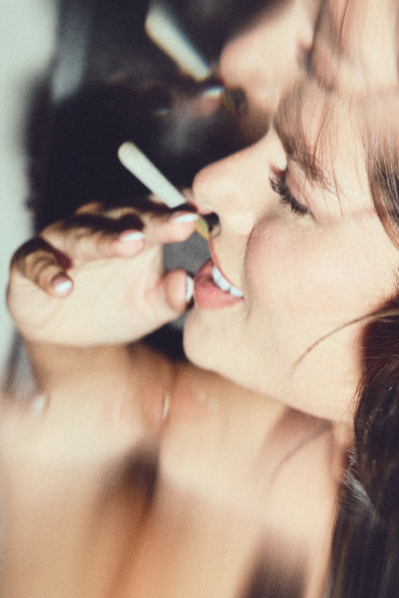 A woman with dark hair and fair skin is close to her face, holding a cigarette near her lips, with her eyes closed.