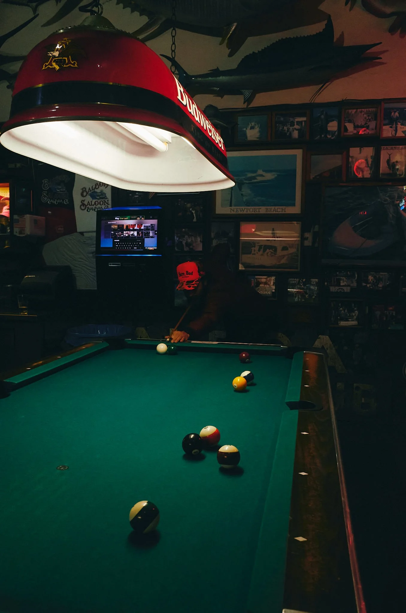 A pool table in a dimly lit bar with several billiard balls on the green felt, a pool cue, framed pictures on the wall, a computer monitor, and a vintage Budweiser lamp hanging above.