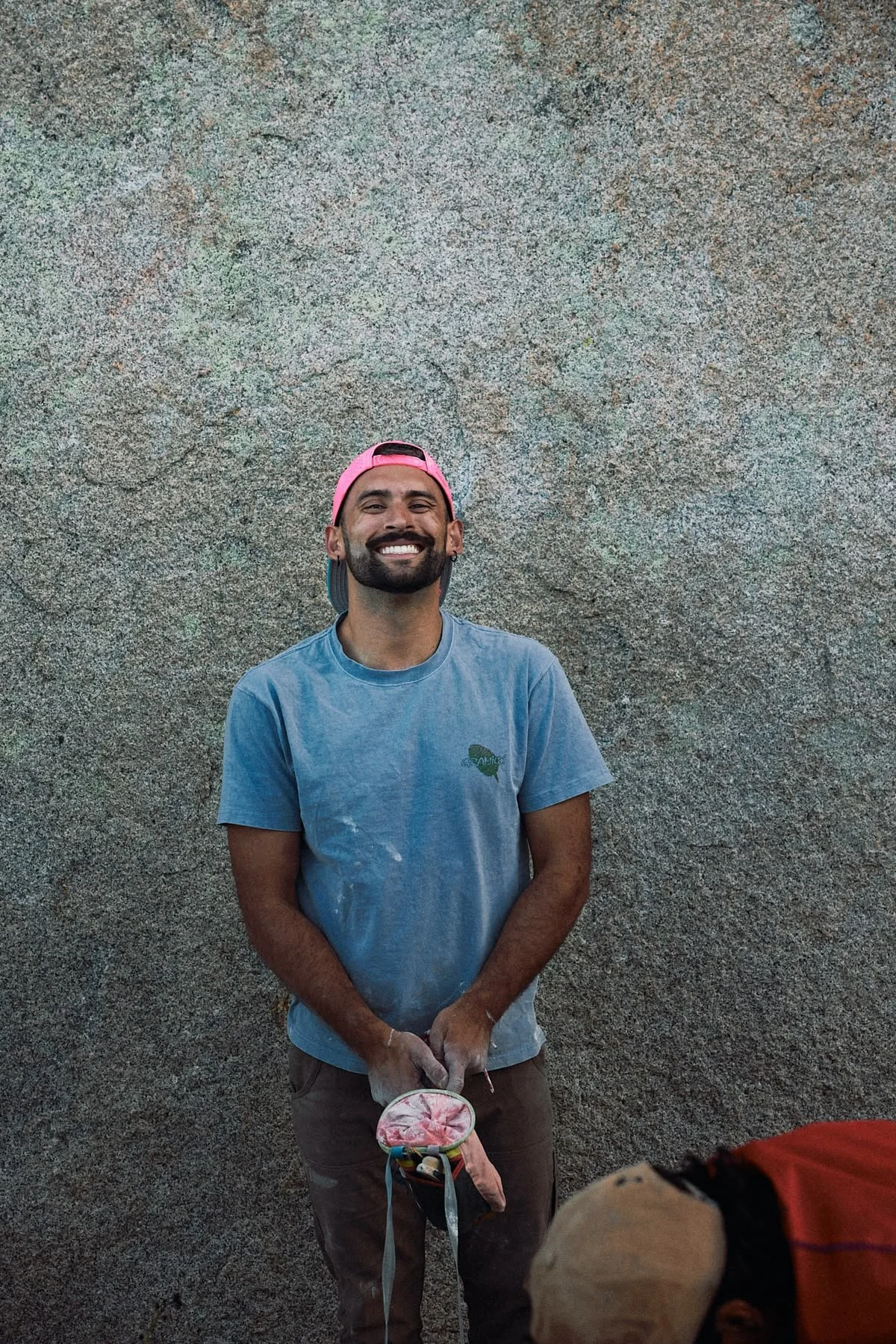A man with a beard and mustache wearing a pink backwards cap, blue t-shirt, and brown pants standing in front of a large textured rock wall, smiling at the camera.