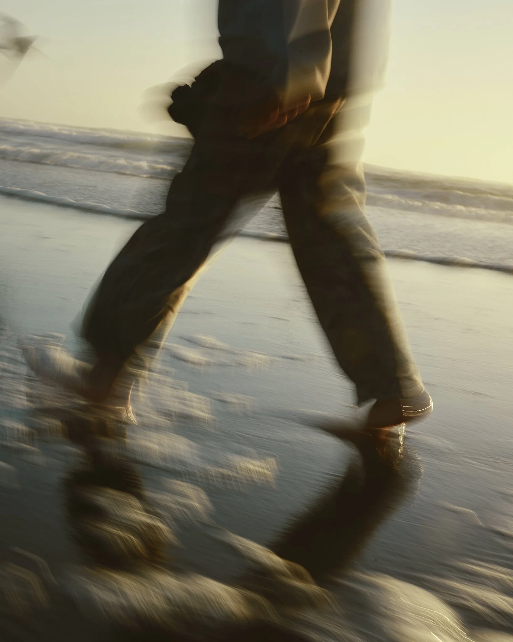 A person walking along the shoreline at sunset with waves crashing at their feet, captured with a motion blur effect.