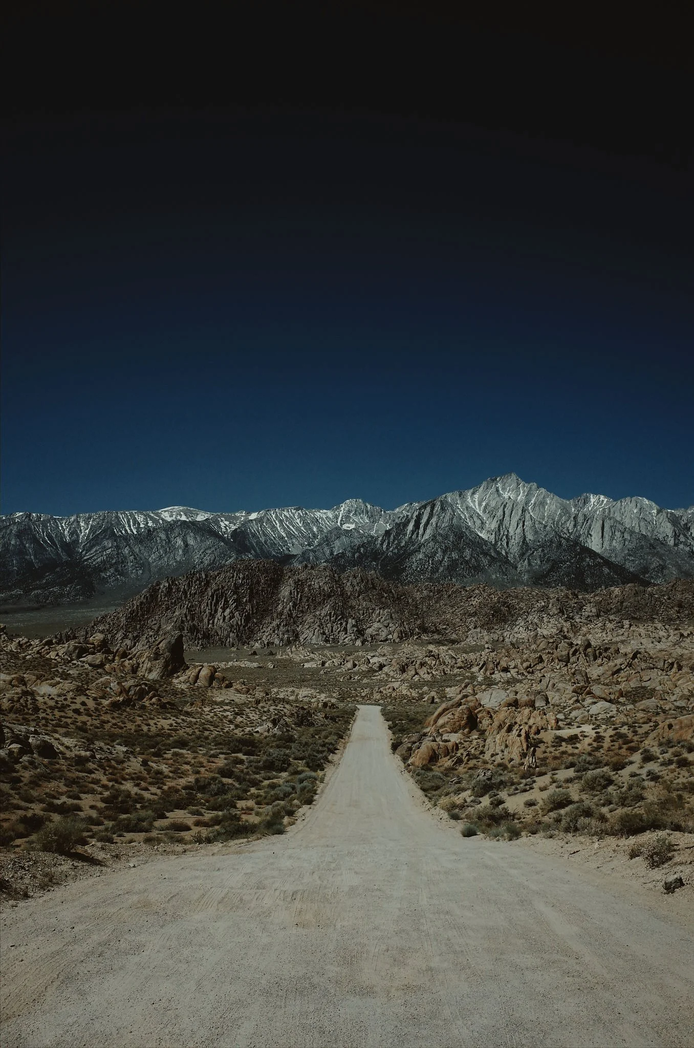 A dirt road stretching through a rocky desert towards snow-capped mountains under a dark blue sky.