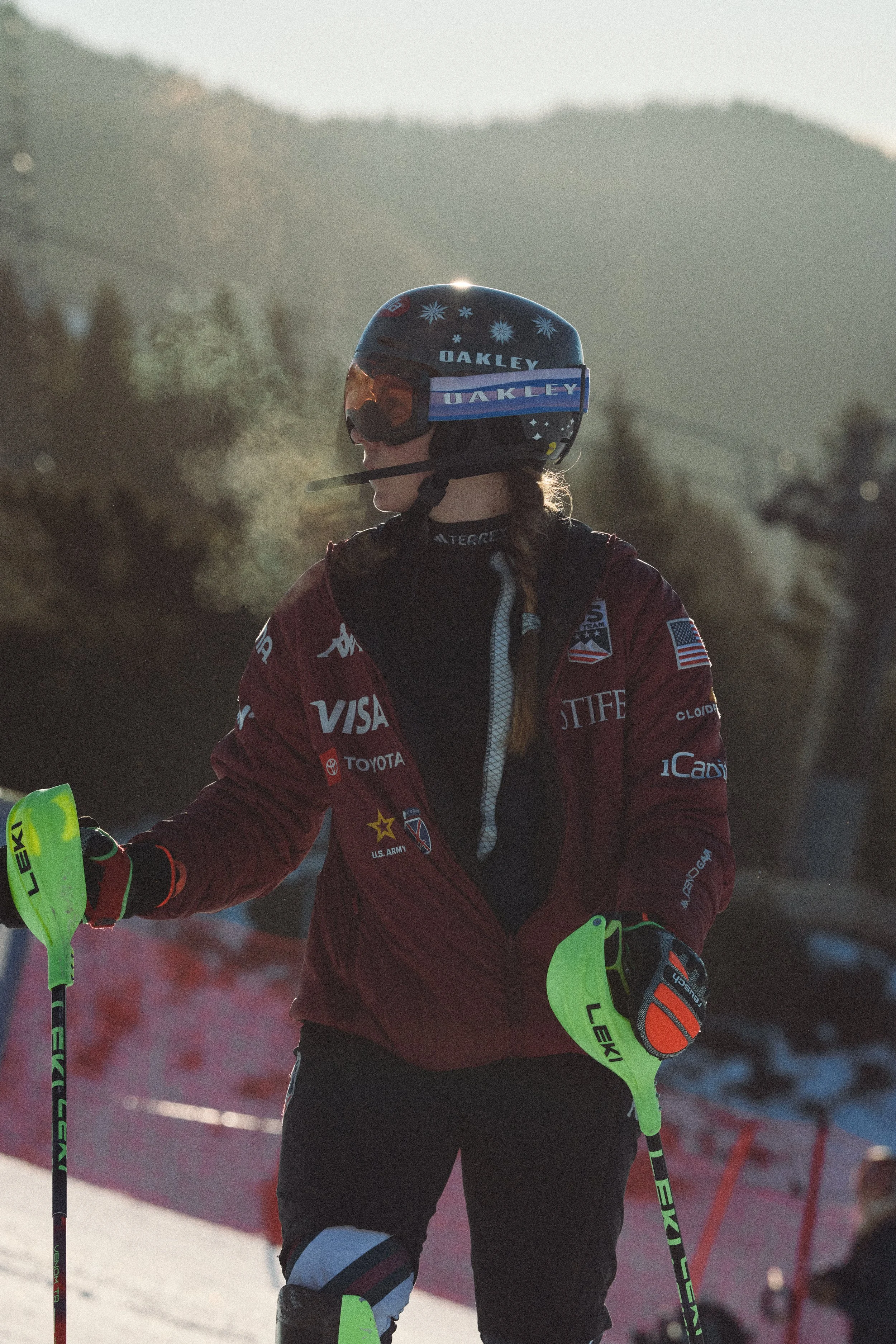 A skier in a red jacket with sponsor logos, wearing a helmet and goggles, holding ski poles on a snowy mountain with a mountainous landscape in the background.
