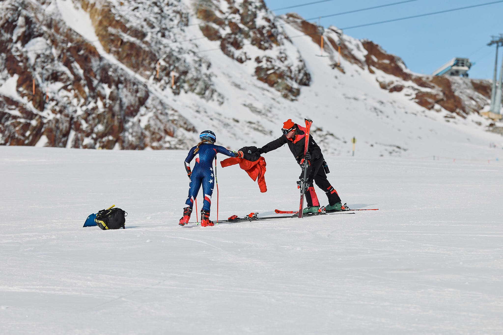 A ski instructor helping a young female skier on a snowy slope in a mountainous area with ski lifts and a chairlift in the background.