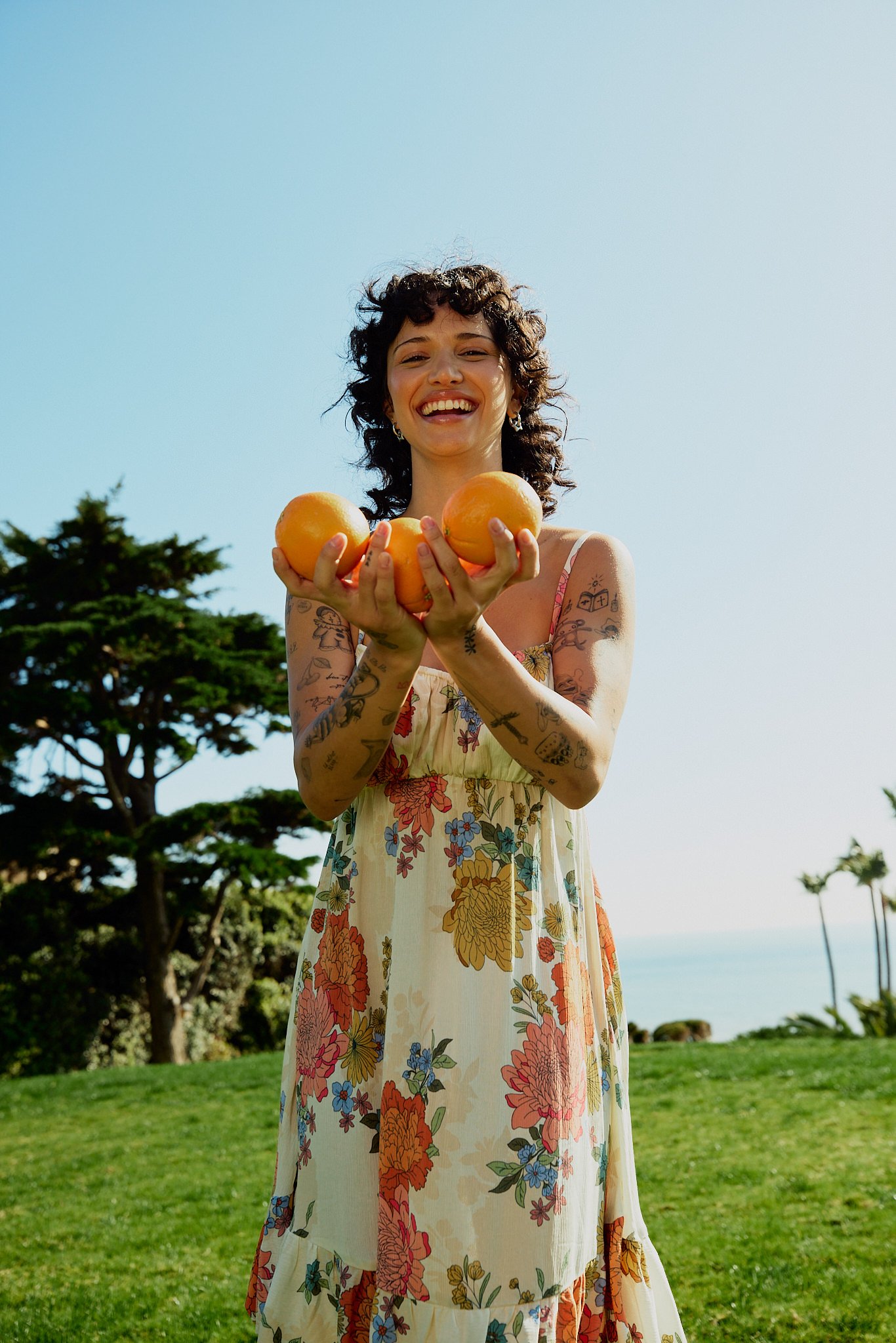 A woman with short curly hair smiling and holding four oranges, standing outdoors on a sunny day with green grass, trees, and a clear blue sky in the background.