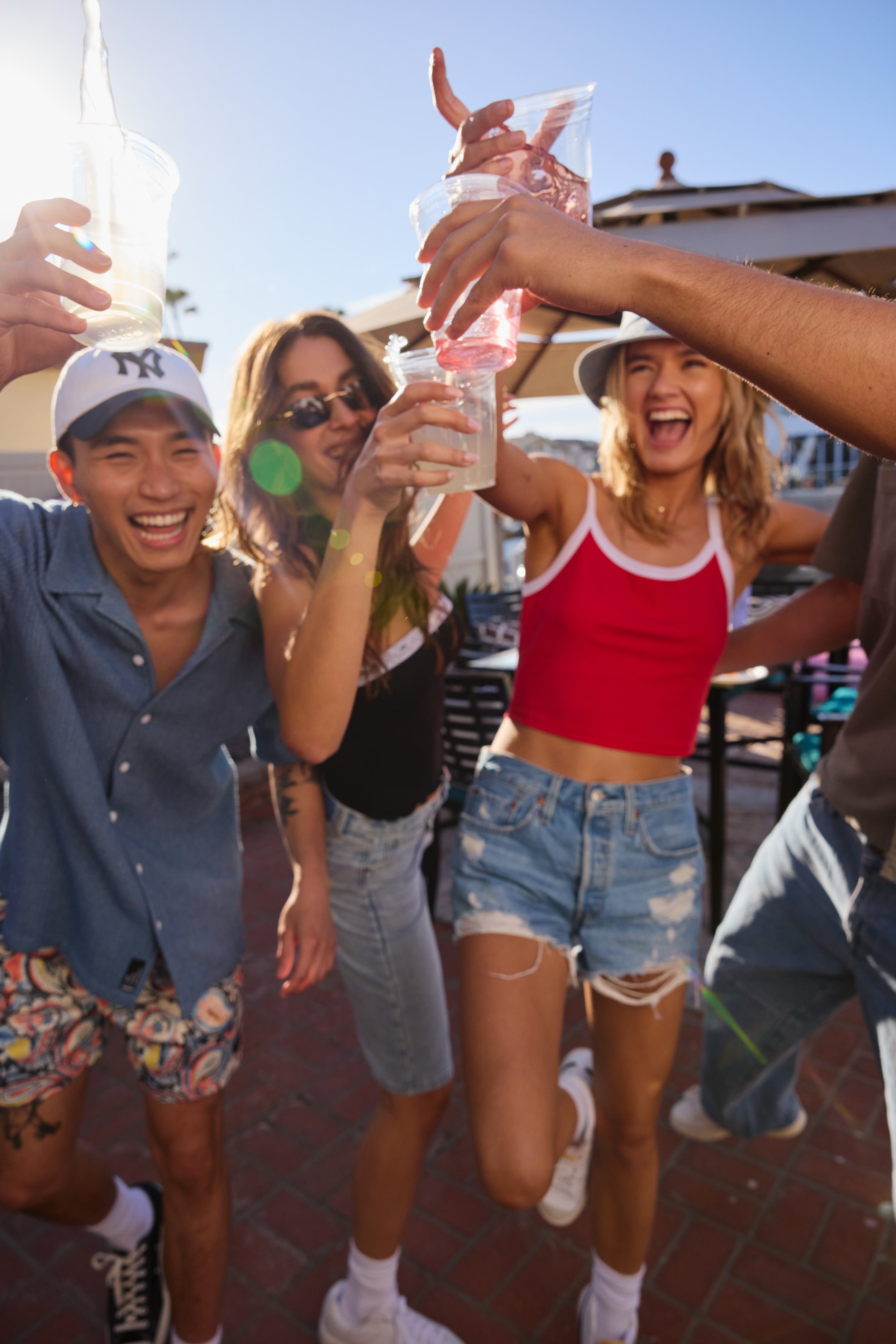 A group of young friends celebrating and making a toast outdoors on a sunny day, with some wearing sunglasses and casual summer clothing.