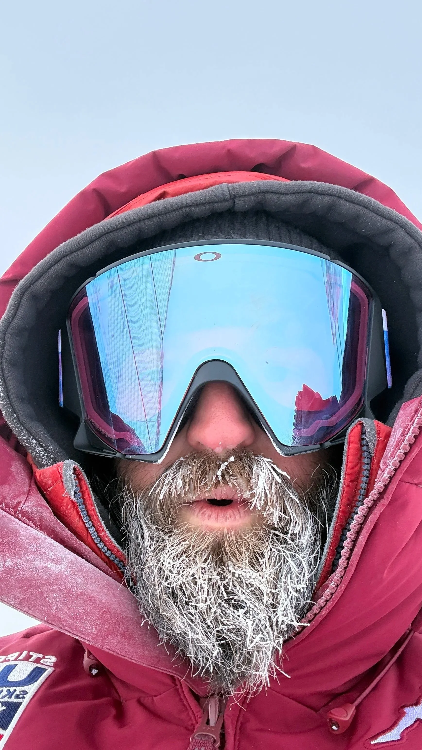 A person wearing a red winter jacket and goggles with a reflective blue lens, head tilted downward, with a white beard and some ice or snow on the beard and jacket, against a light sky background.