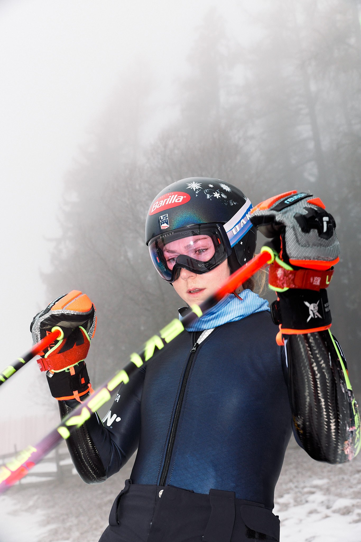 A woman dressed in winter gear, including gloves, a helmet, and a jacket, holding ski poles in a snowy environment with trees in the background.