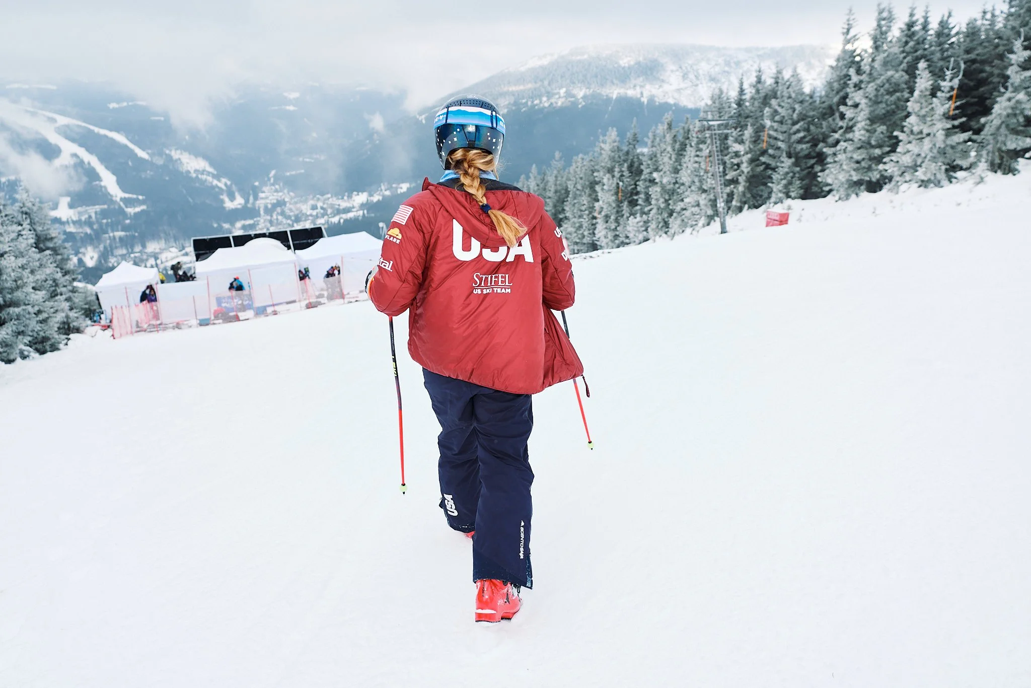 A female skier wearing a red USA jacket, black ski pants, and pink ski boots walking in a snowy mountain landscape with trees and ski lifts in the background.
