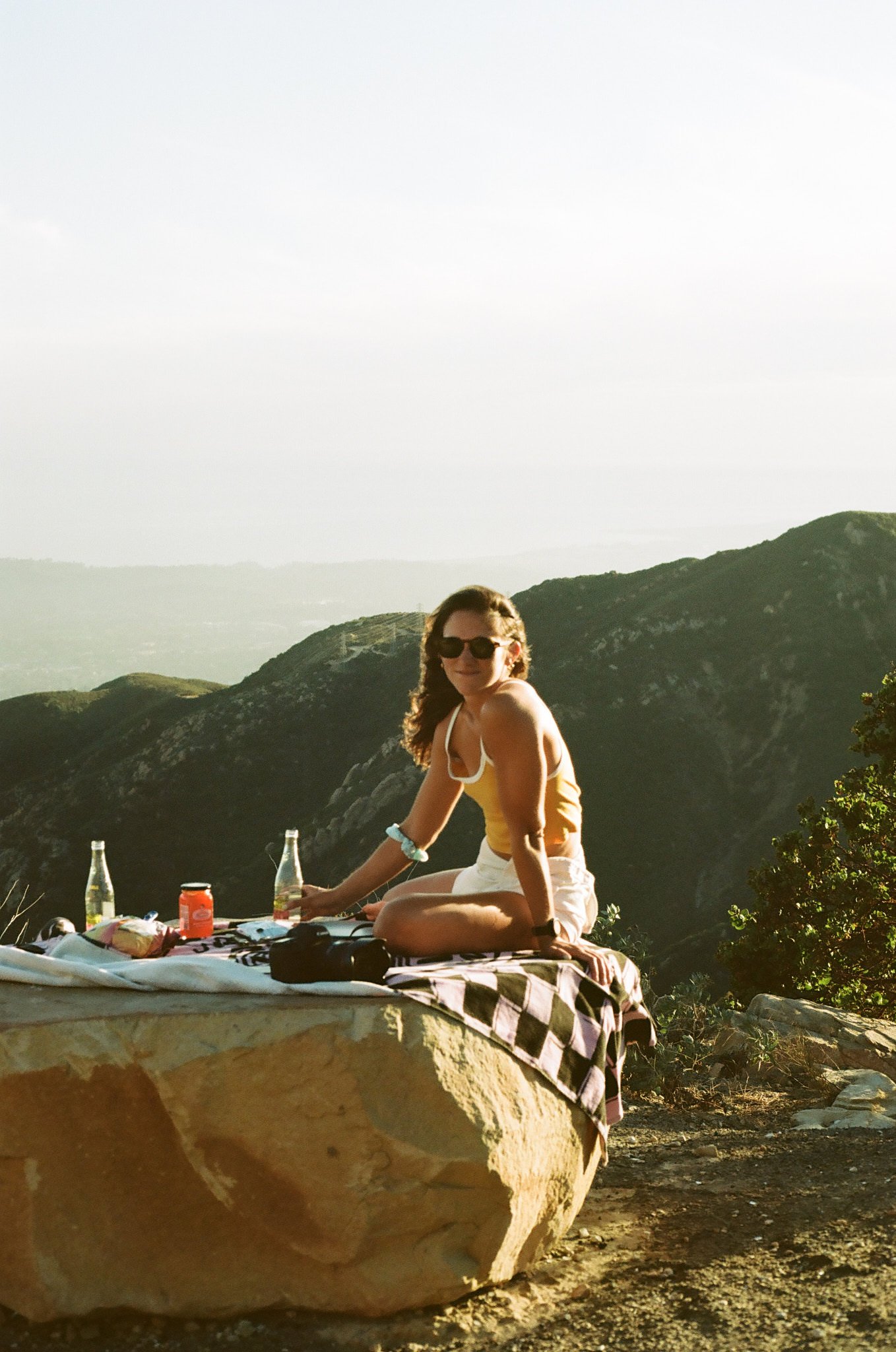 A woman sitting on a blanket outdoors with a mountain landscape in the background, surrounded by drinks and snacks.