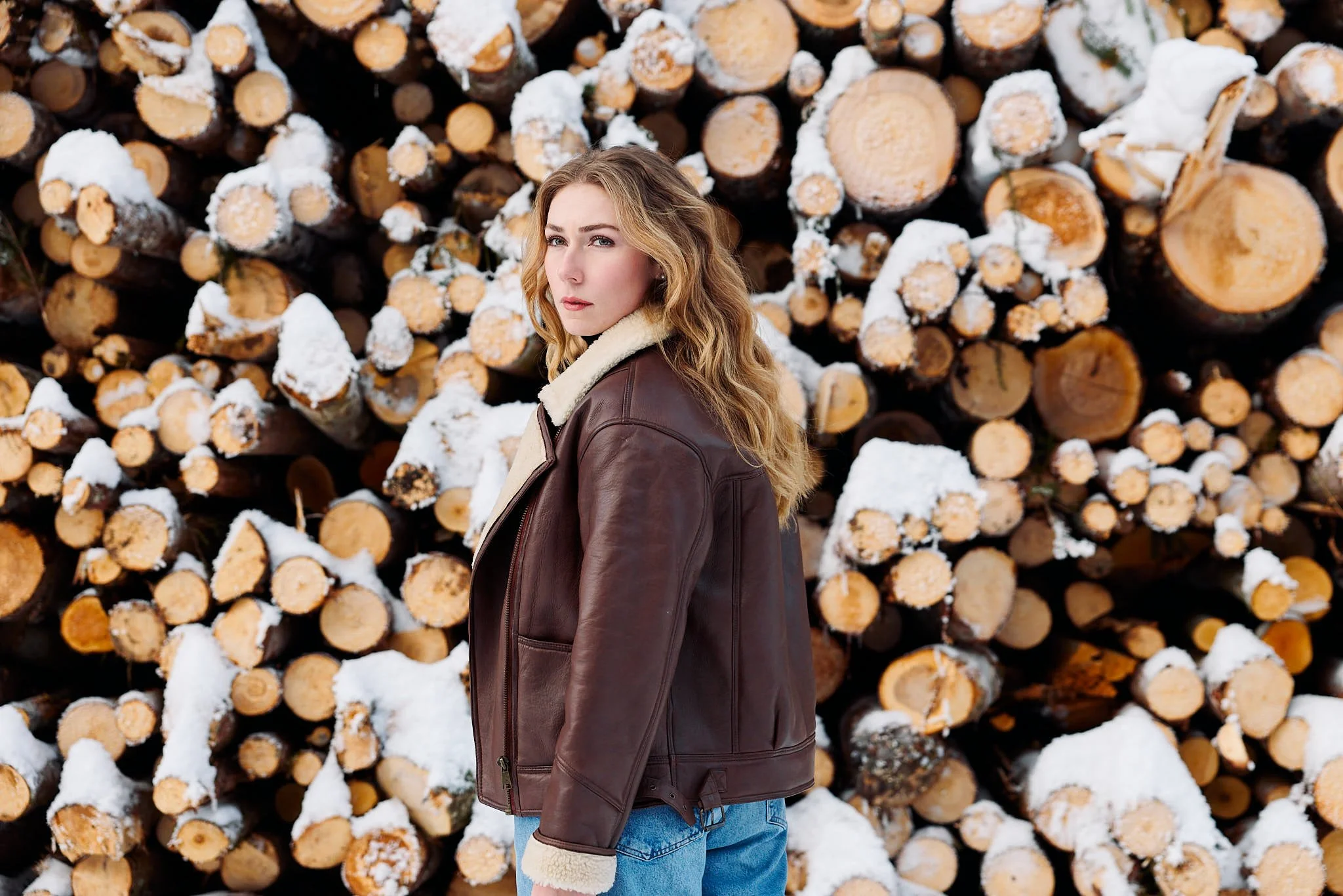 A woman with long wavy blonde hair, wearing a brown leather jacket with a shear lining and light blue jeans, standing in front of a large stack of cut logs covered in snow.