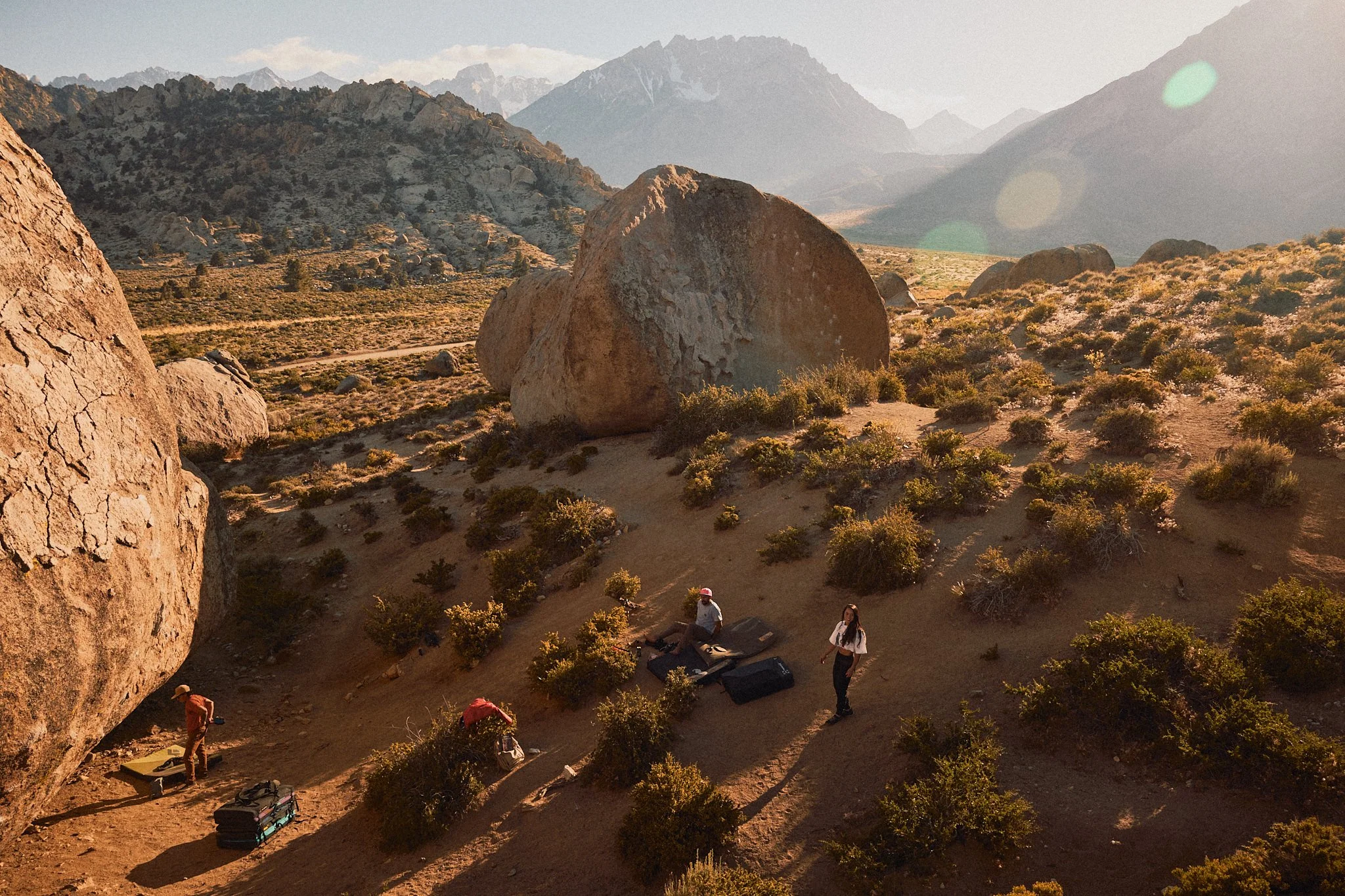Hikers setting up gear in a mountainous desert landscape with large boulders, low shrubs, and distant snow-capped peaks under a clear sky.