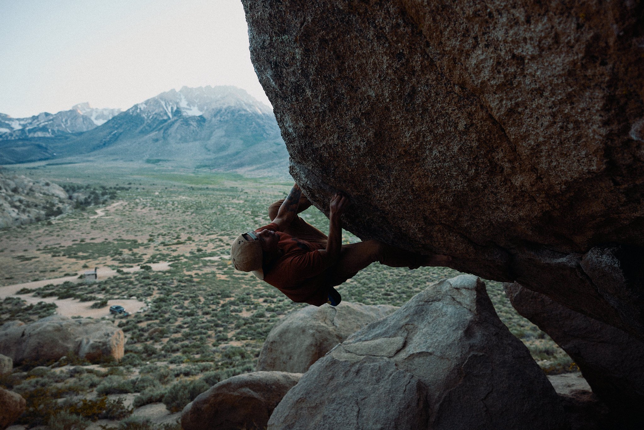 A person wearing a helmet rock climbing on a large boulder outdoors with mountains in the background.