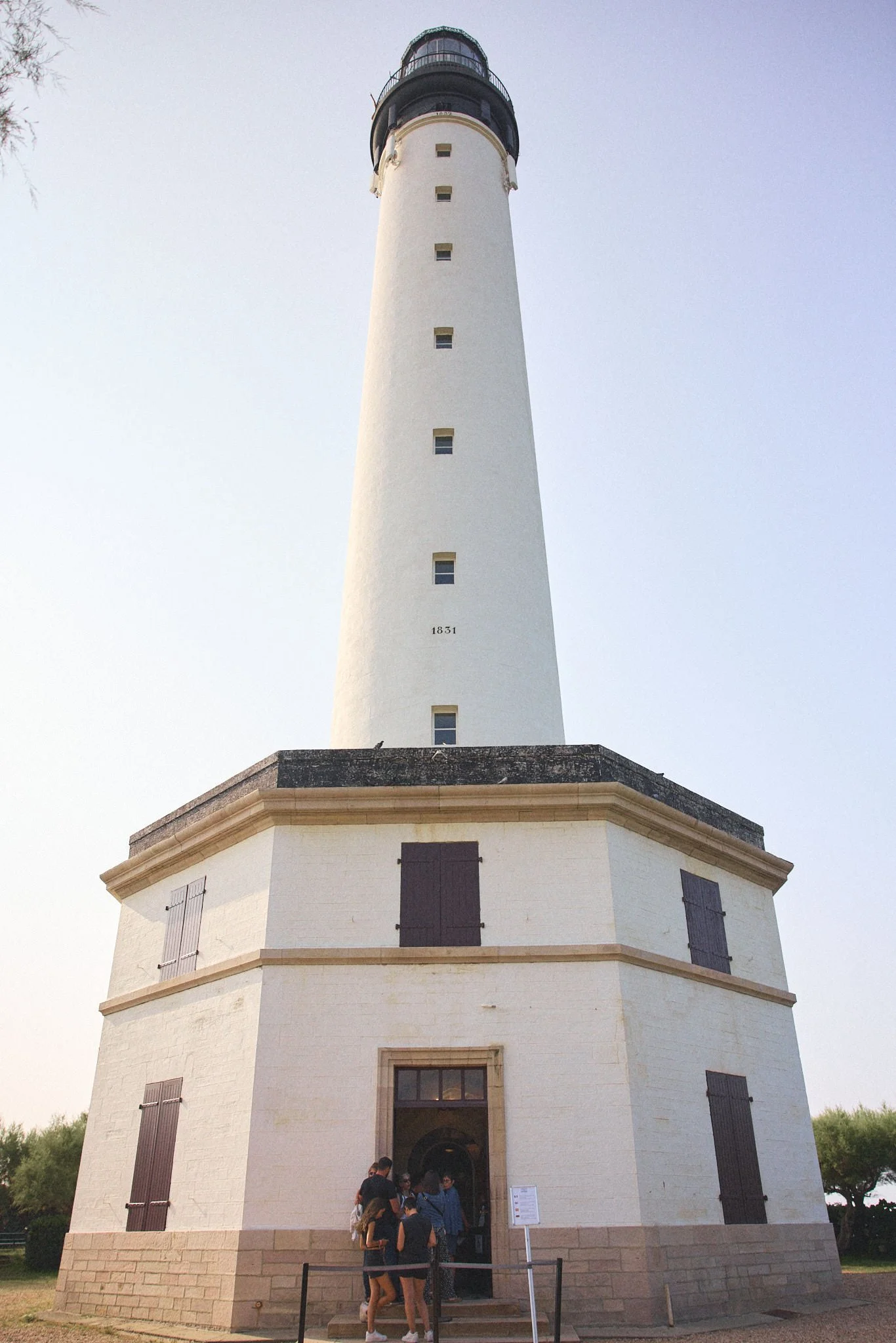 A tall white lighthouse with black trim, surrounded by a group of people, set against a clear sky.