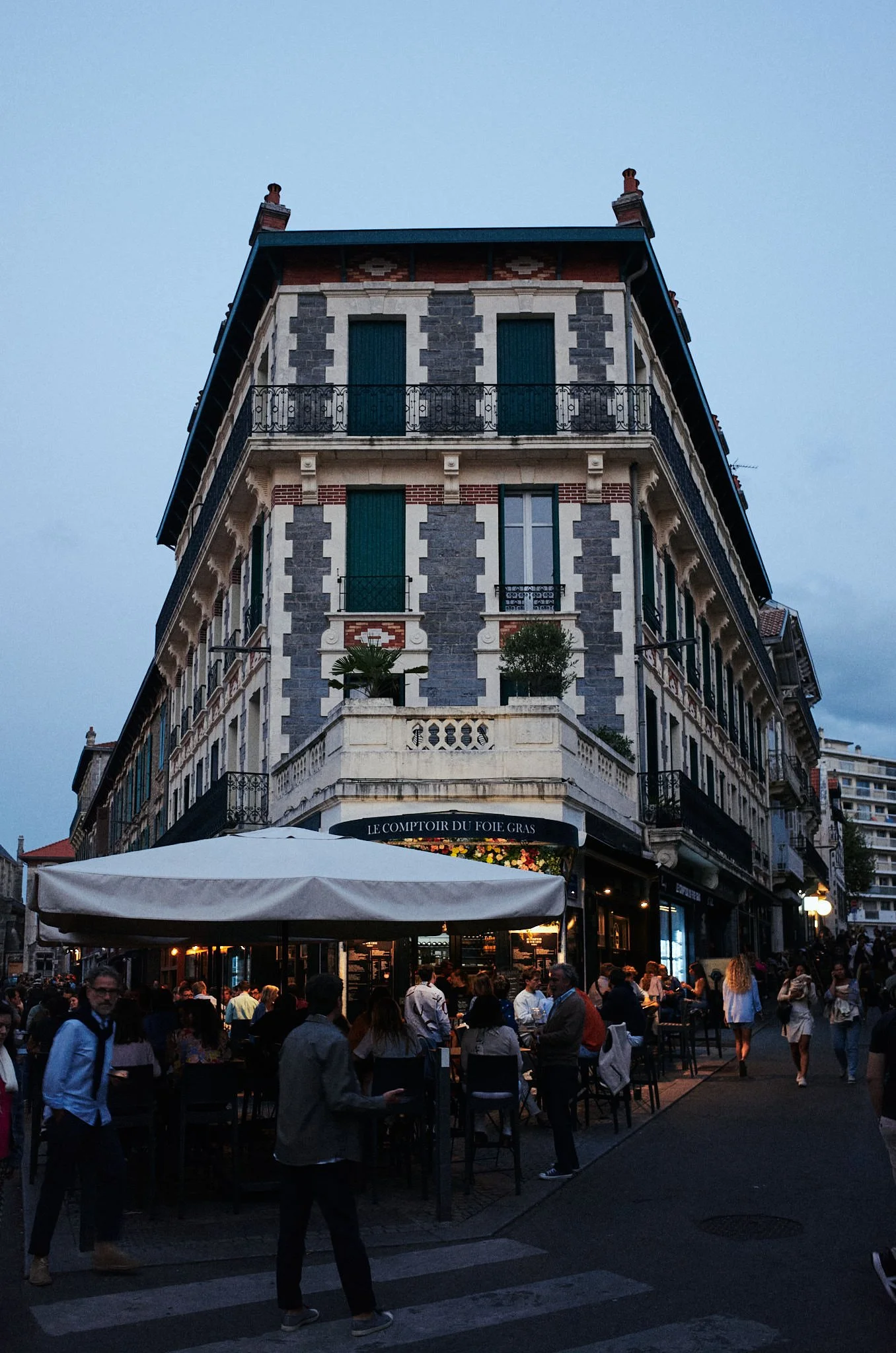 A bustling outdoor café with tables and people dining, situated on a city street corner beneath a large, elegant corner building with green shutters and wrought iron balconies, during dusk.
