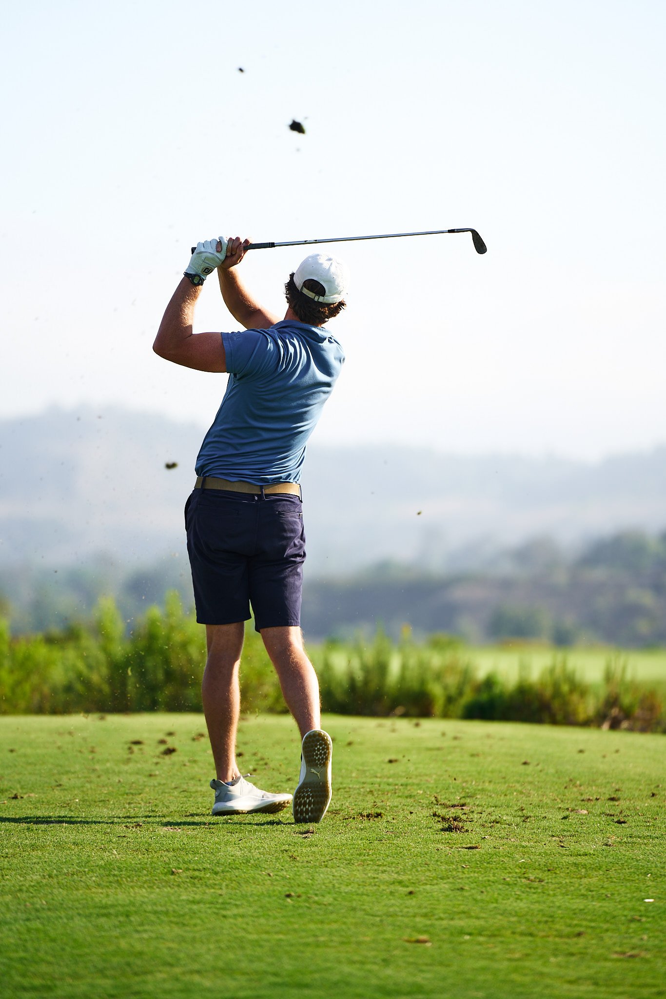 Man in a blue shirt and navy shorts swinging a golf club on a golf course with a bright sky and lush green landscape.
