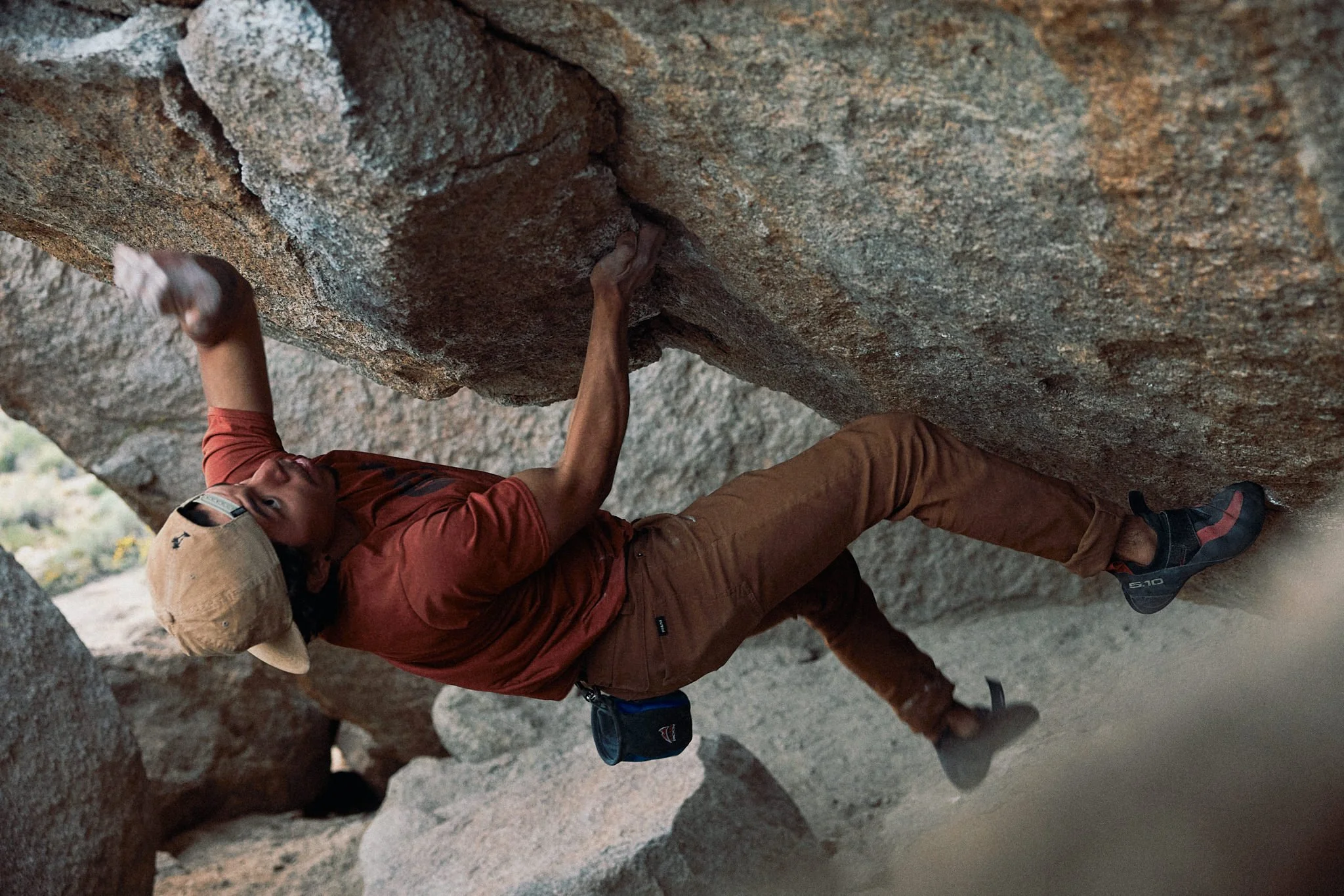 A man wearing a hat and climbing gear is rock climbing, reaching for a handhold on a granite cliff face.