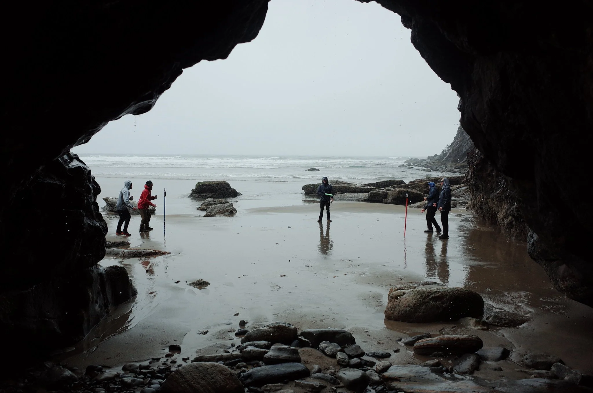 View from inside a dark cave looking out to the beach where five people are walking and standing near the shoreline, with rocks and the ocean visible in the background on a cloudy day.