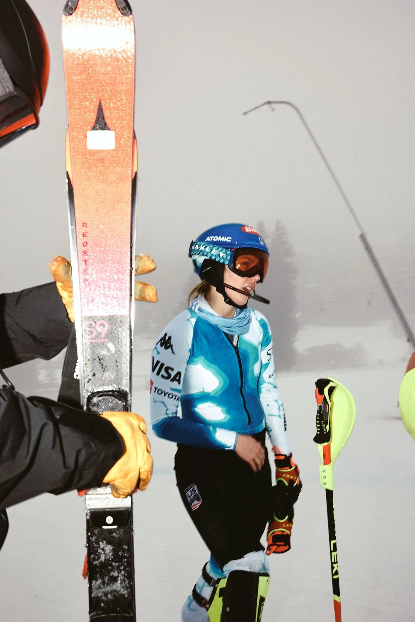 A female skier wearing a blue helmet and ski gear holds a yellow ski in a snowy environment, with a ski in the foreground and trees in the background.