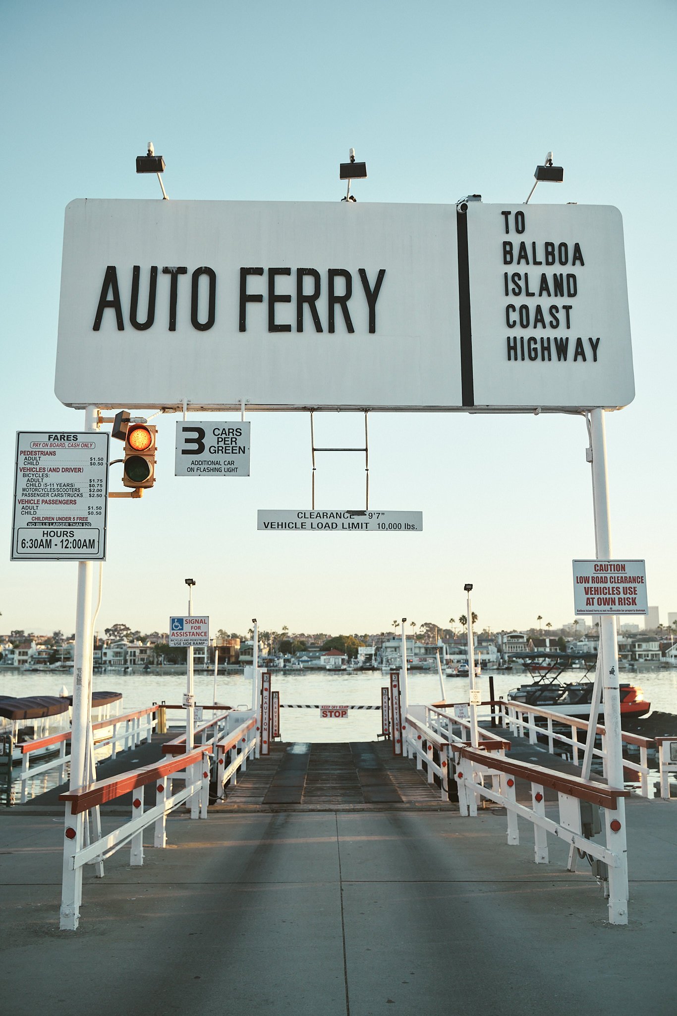 A ferry dock with a sign that reads 'Auto Ferry' and directional signs to Balboa Island Coast Highway, a boat in the water, and residential buildings in the background, under a clear sky.