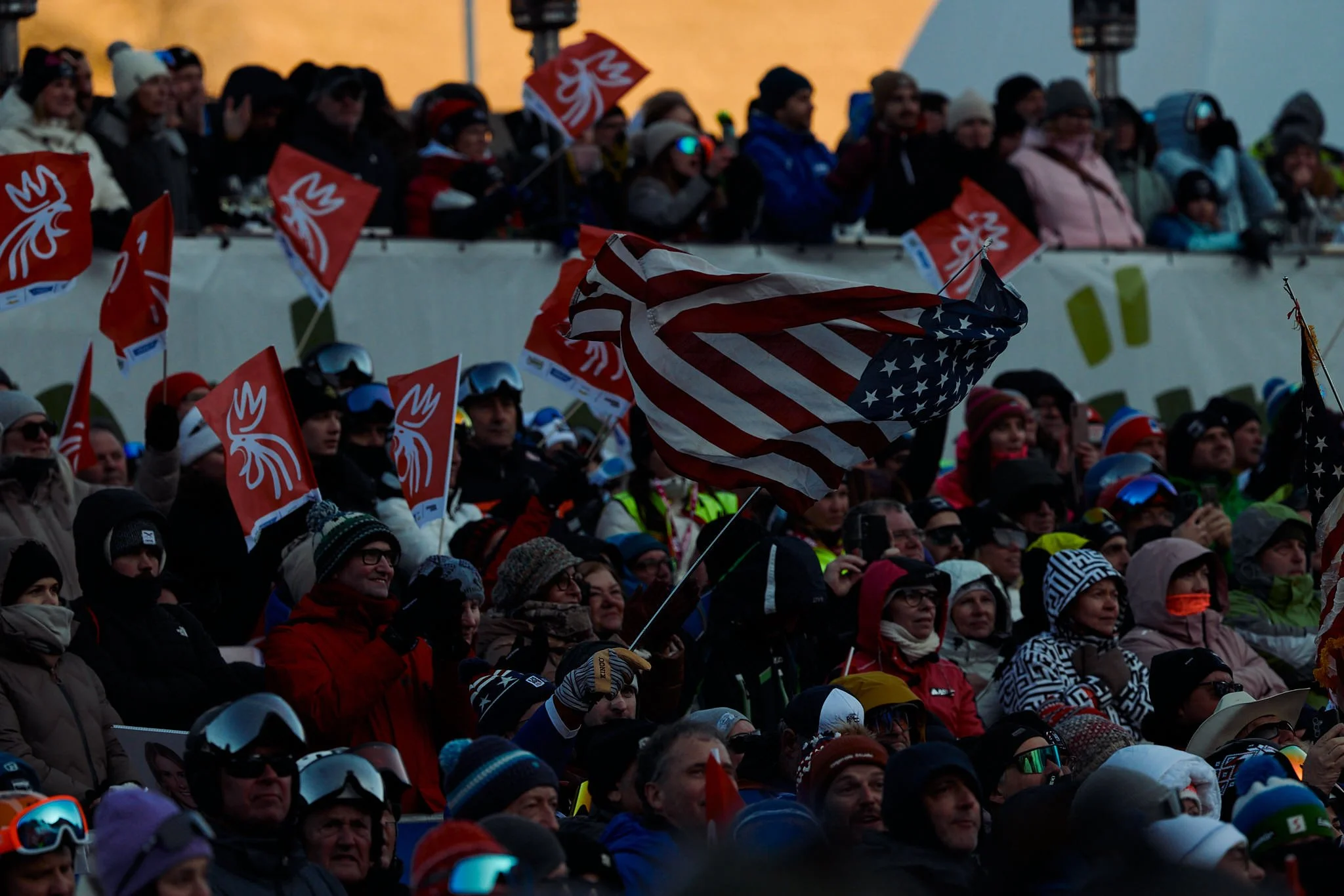 A large crowd of people bundled up in winter clothing, some wearing helmets and goggles, gathered outdoors during what appears to be a winter sports event. Many spectators are waving red flags with a white emblem and an American flag.