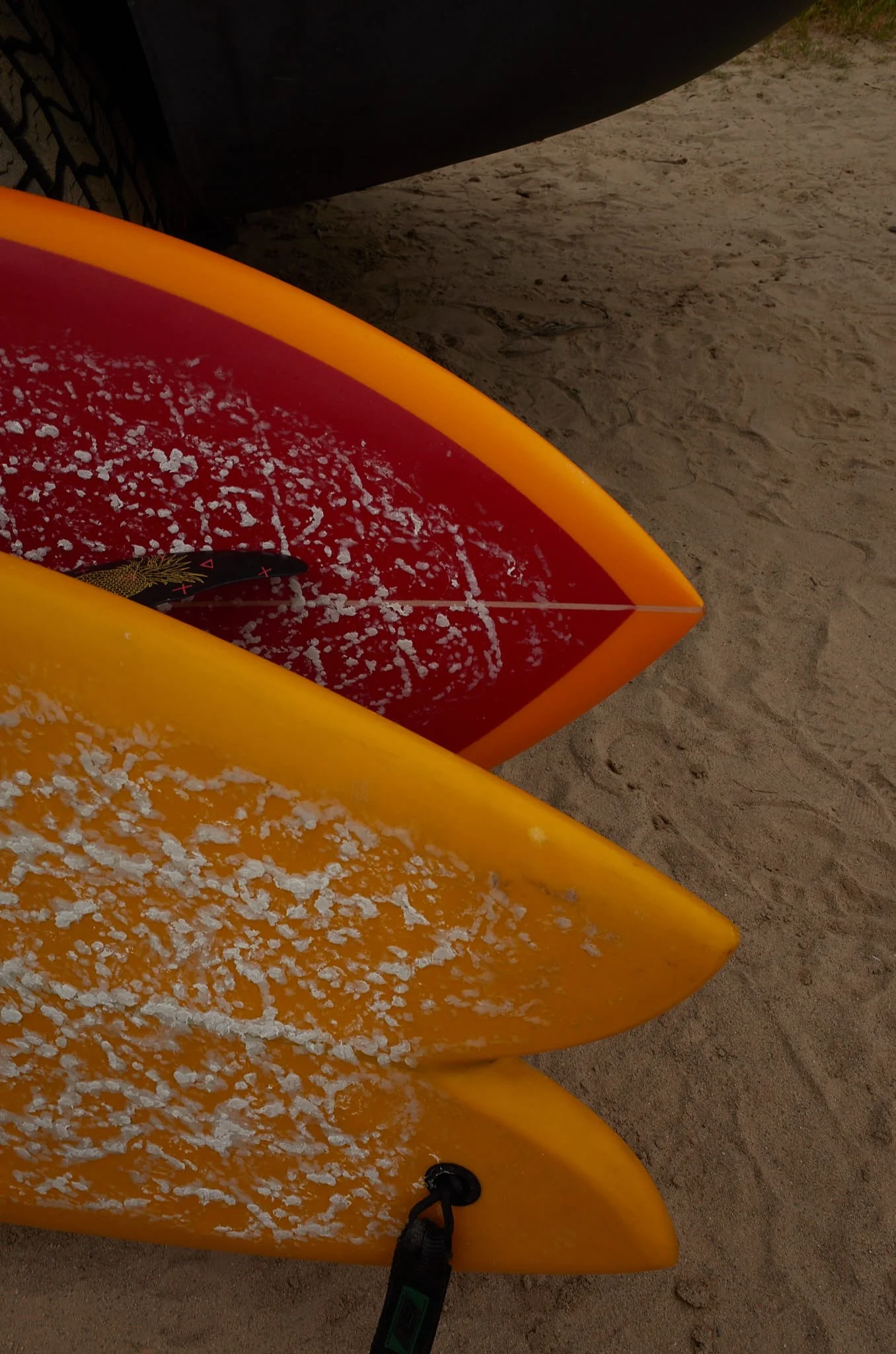 Close-up of a yellow surfboard with white foam remnants resting on sandy beach, a part of a red surfboard is visible underneath.