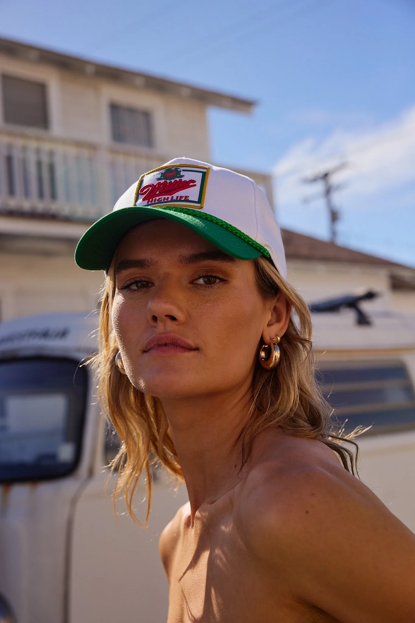 A woman with blond hair wearing gold hoop earrings and a white cap with green trim, standing outdoors on a sunny day, with a white house and blue sky in the background.