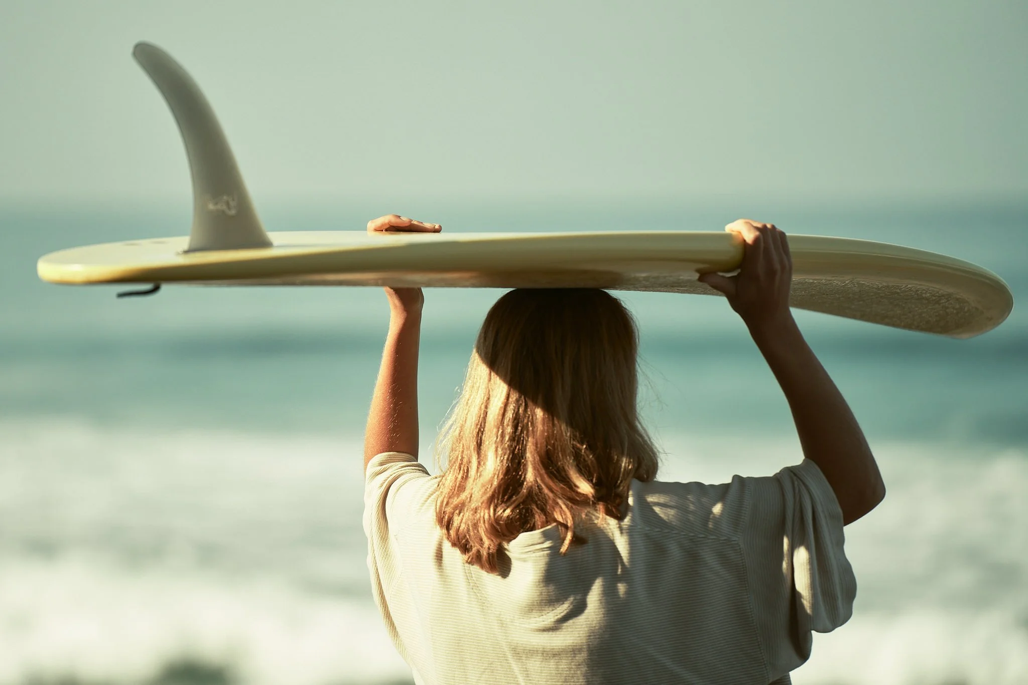 A woman with long brown hair carrying a surfboard on her head at the beach.